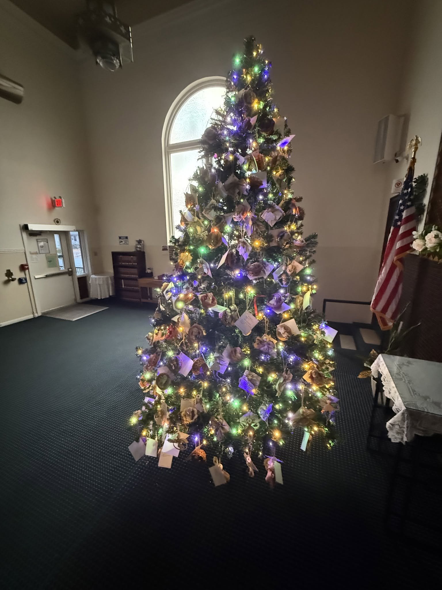 A decorated Christmas tree with colorful lights and ornaments inside a room with a high ceiling and a large arched window in the background.