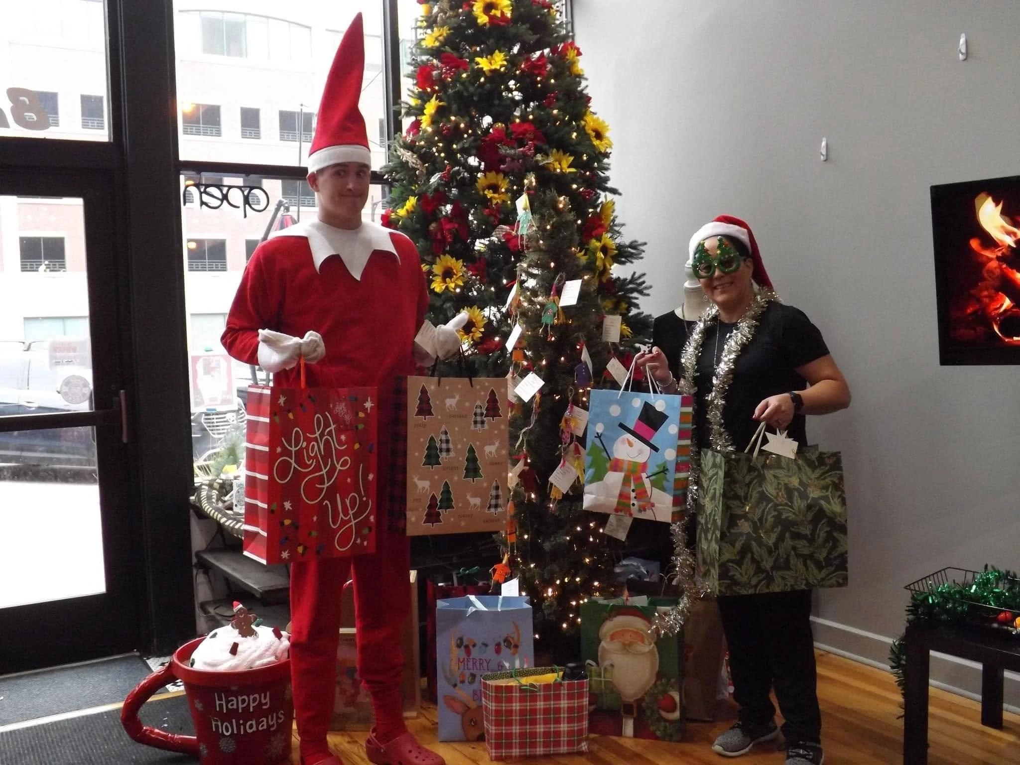 Two people dressed in holiday-themed costumes, one as an elf and the other as Santa's helper, standing near a decorated Christmas tree with gifts around it. The person on the left is wearing a red elf costume with a tall hat, and the person on the right is wearing a Santa hat, green glasses, tinsel, and a T-shirt. They are inside a room with large windows, and there are Christmas bags and a festive display.