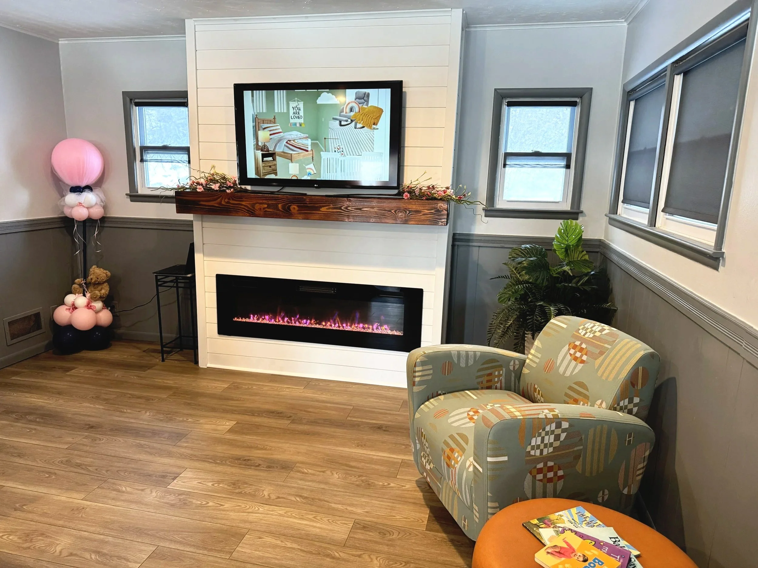 A living room with a fireplace, a pink balloon sculpture in the shape of a bunny on a stand, three small windows, a patterned armchair, and a few books on an orange stool.