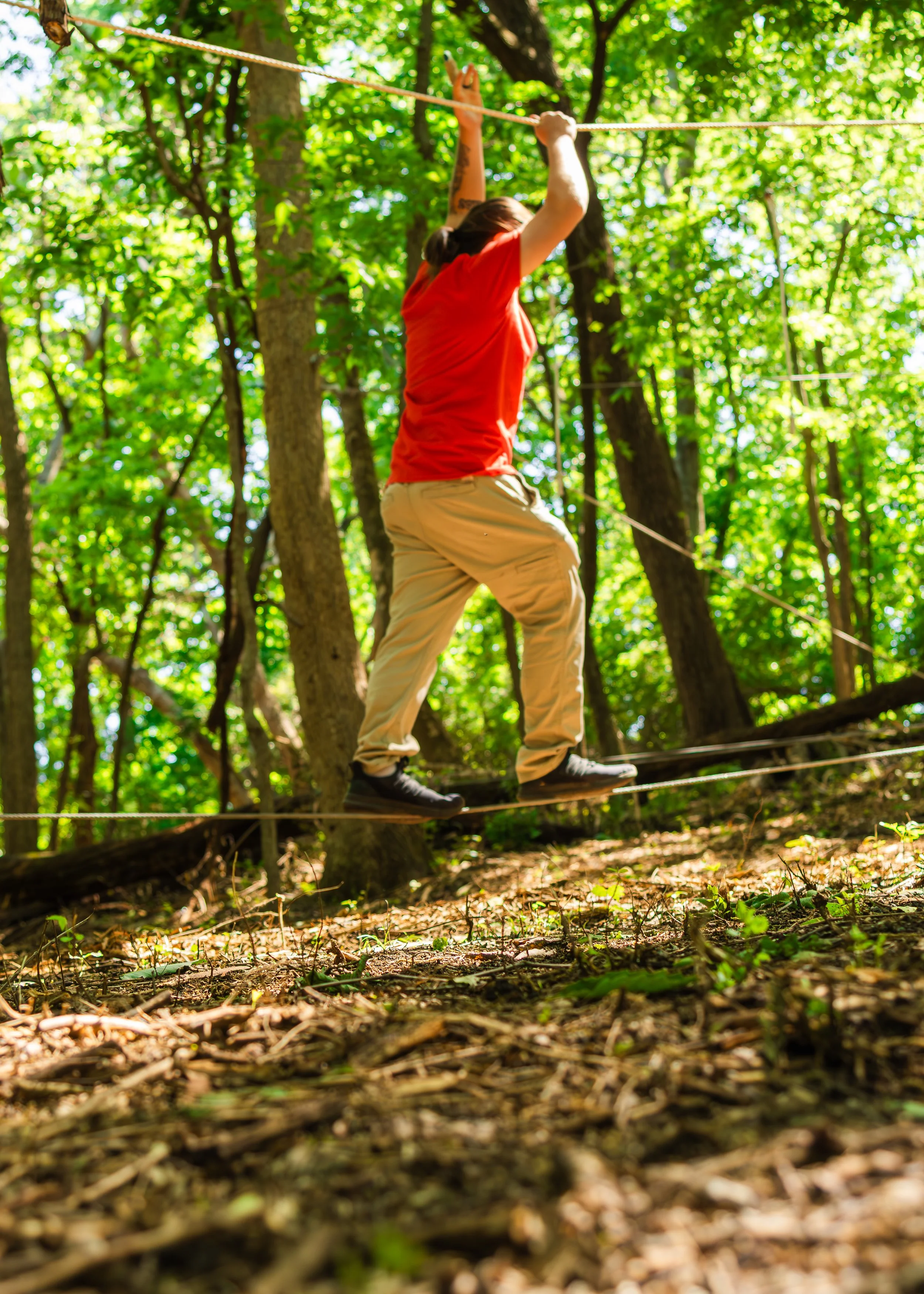 Woman in orange shirt and beige pants walking on a slackline in a forested area with sunlight filtering through green leaves.