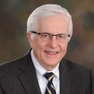 A smiling elderly man with white hair and glasses, wearing a dark suit, white shirt, and striped tie, against a blurred outdoor background.