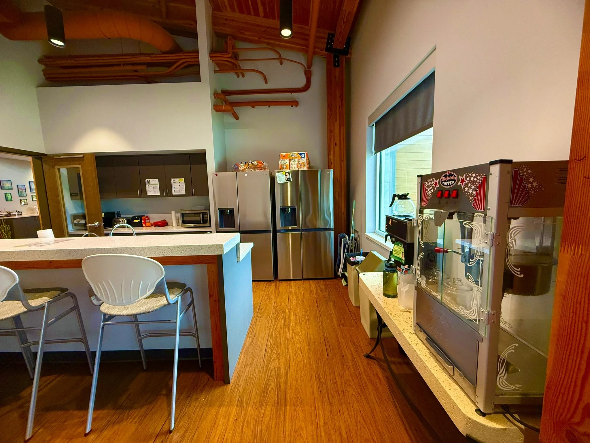 Kitchen area with white island, three chairs, stainless steel refrigerator, microwave, and a window with a shade. Ceiling features wooden beams and exposed pipes.