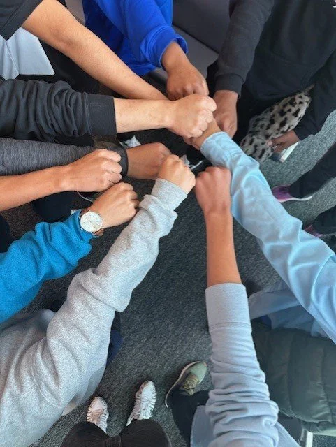 Multiple people in a circle extending their fists together in a show of teamwork.