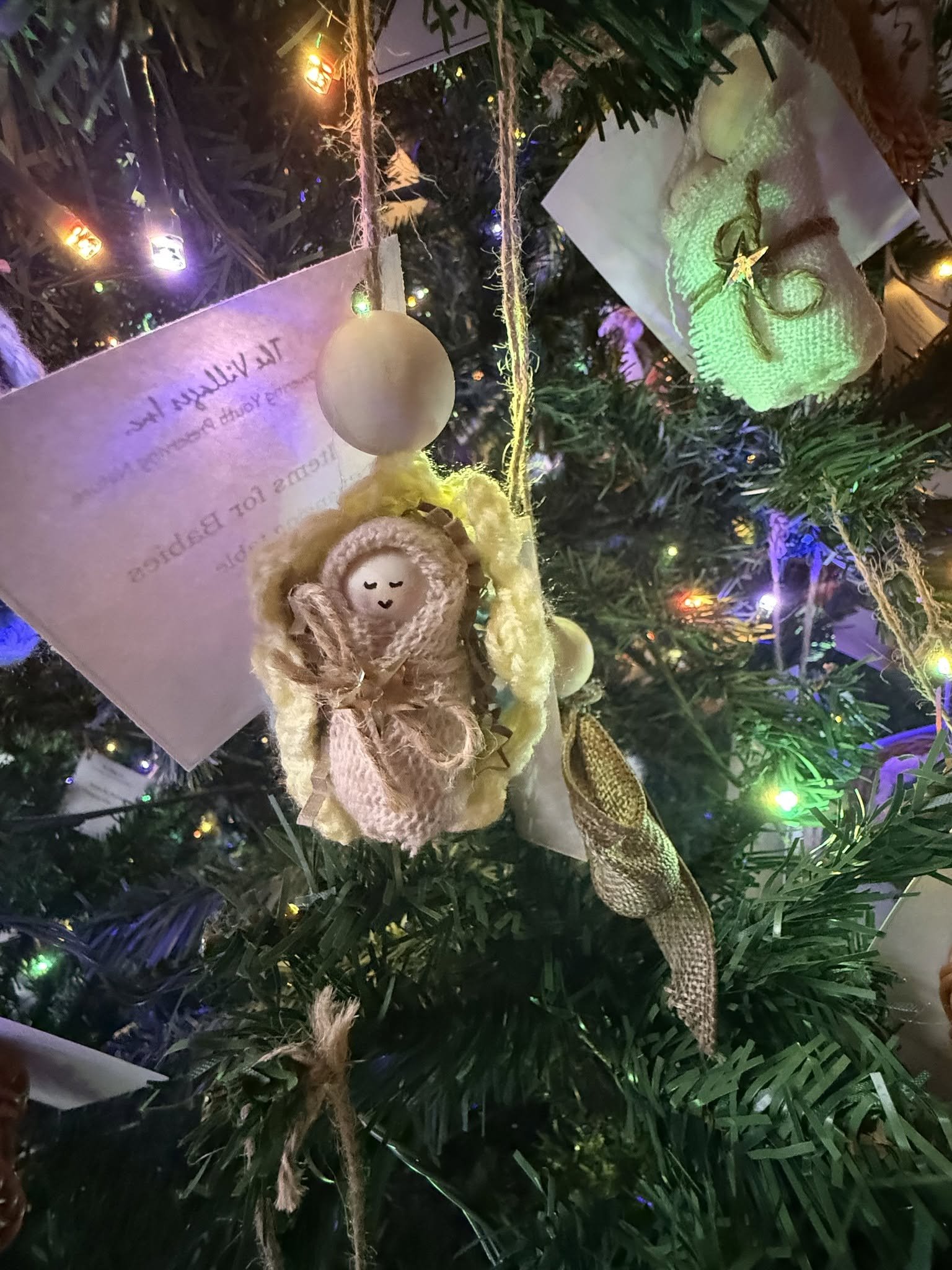 Close-up of a decorated Christmas tree with ornaments, including a small plush angel, a pearl-like ball, ribbons, and multicolored lights.