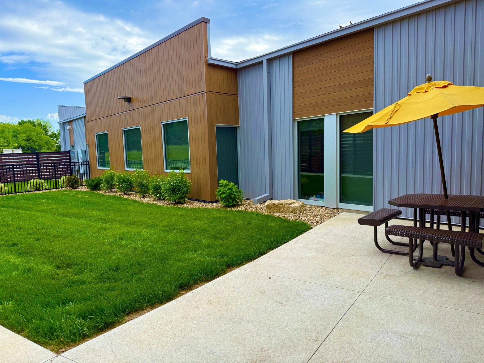 Modern building with a mix of wooden and metal exterior walls, large glass windows, a landscaped yard with green grass and plants, a concrete patio, and a yellow patio umbrella.