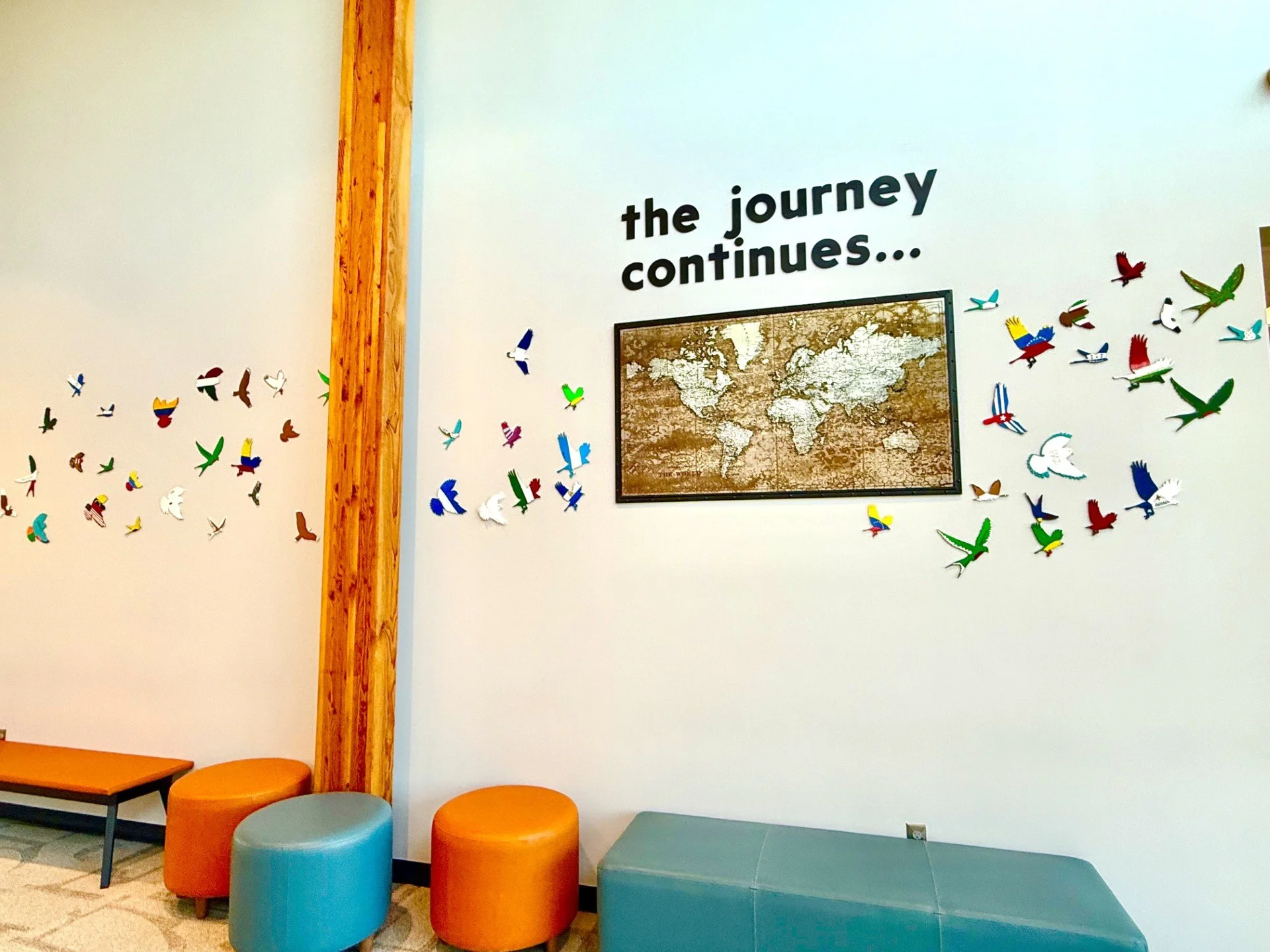 Waiting area with colorful stools, a wooden beam, a world map on the wall, and decorative birds, with the phrase 'the journey continues...'