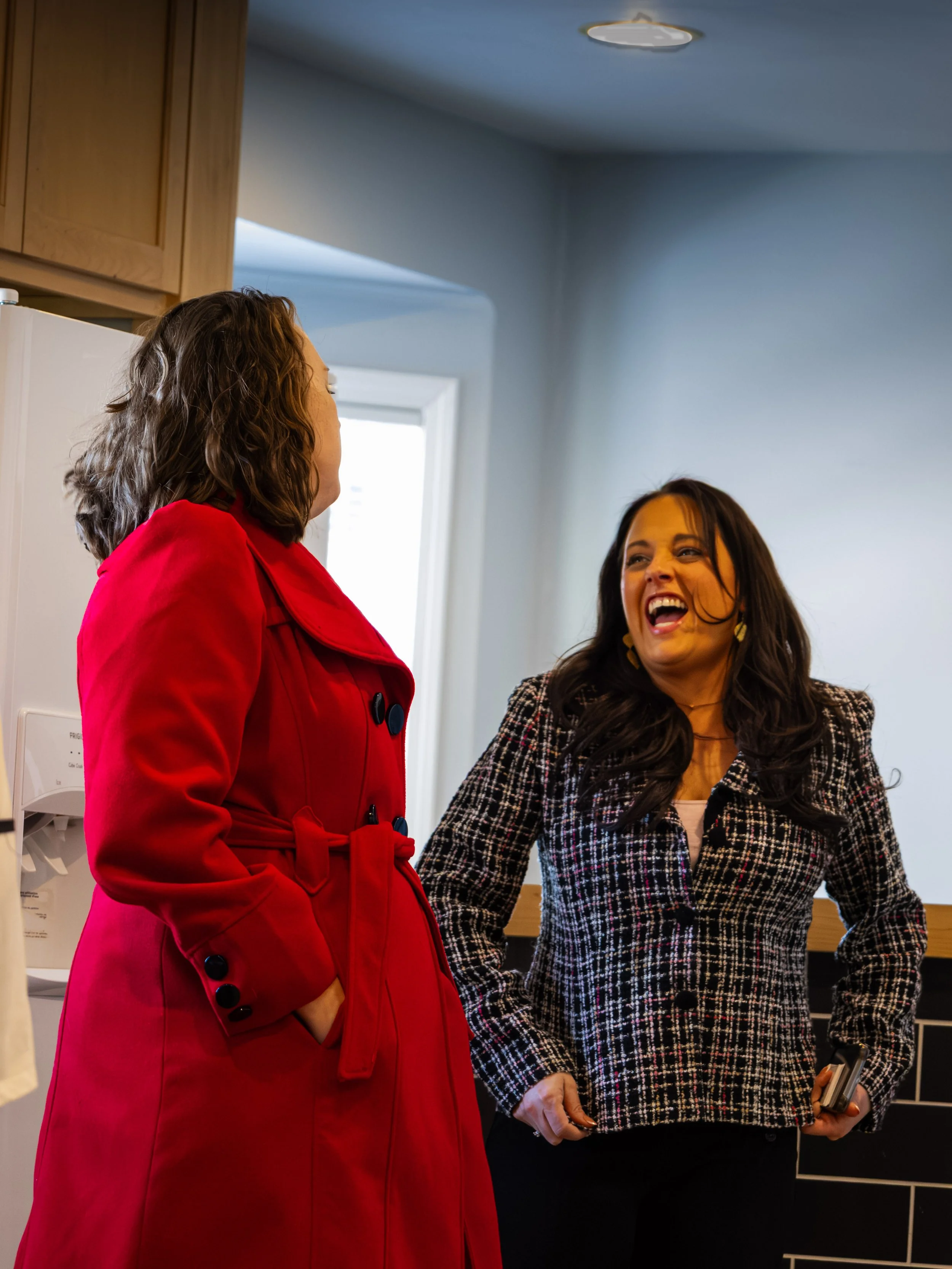 Two women engaging in conversation inside a kitchen. One woman is wearing a bright red coat and the other is in a black and white checkered blazer, laughing and holding a phone.