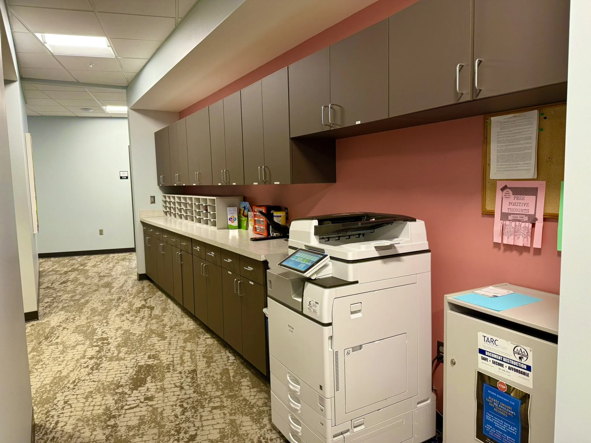 Office copier machine next to a beige filing cabinet, with wall-mounted cabinets above and a bulletin board on the pink wall.