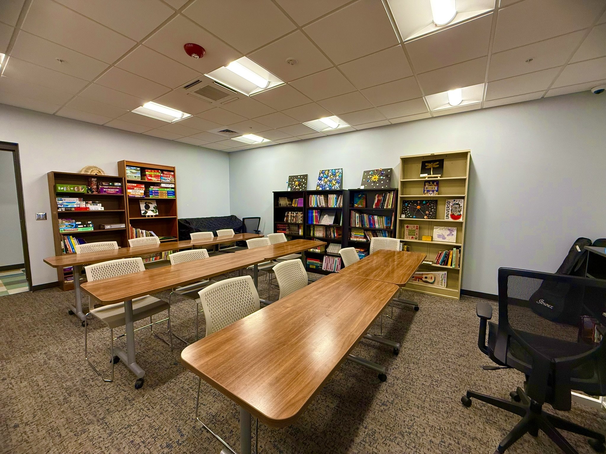 A conference room or classroom with a long wooden table surrounded by white chairs, bookshelves filled with books and art, and a ceiling with fluorescent lighting.