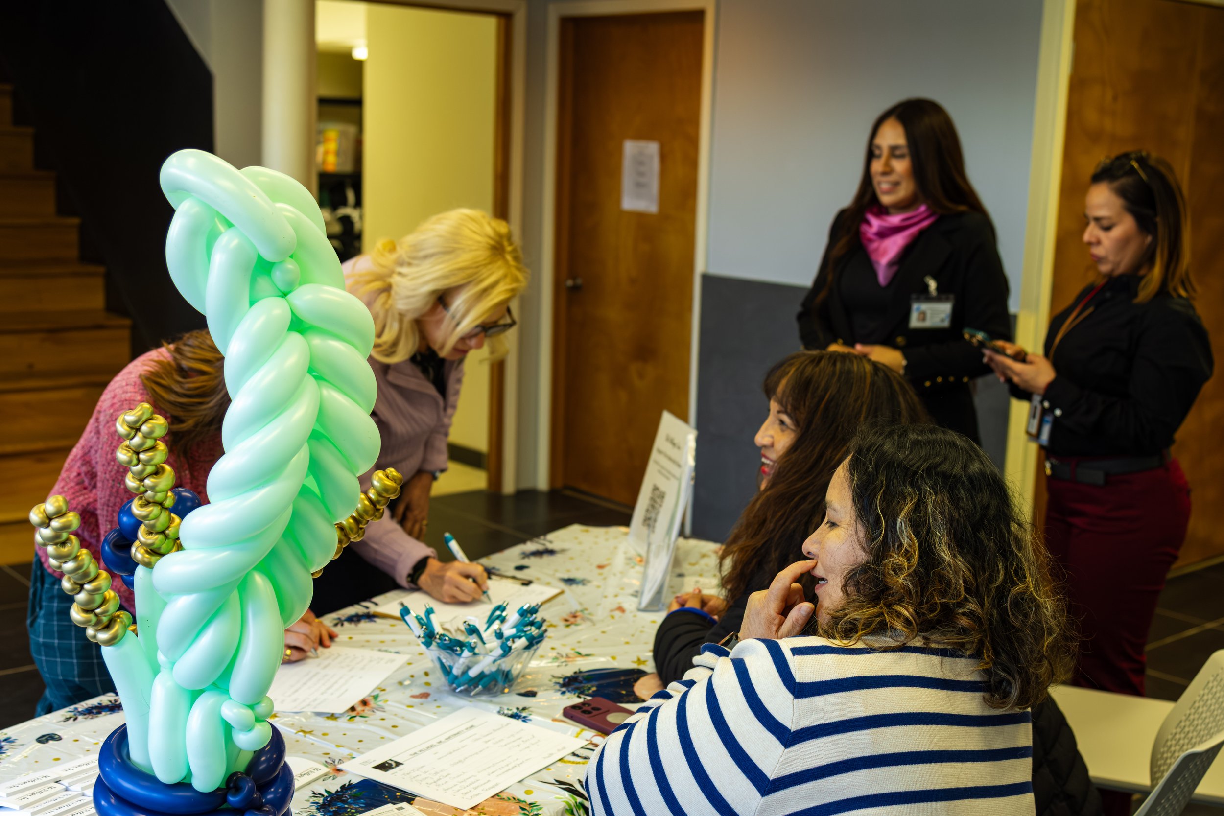 A group of women at a registration or check-in table, with one woman signing in and others standing around, decorated with a large, twisted balloon sculpture in green, light green, navy blue, and metallic gold balloons.
