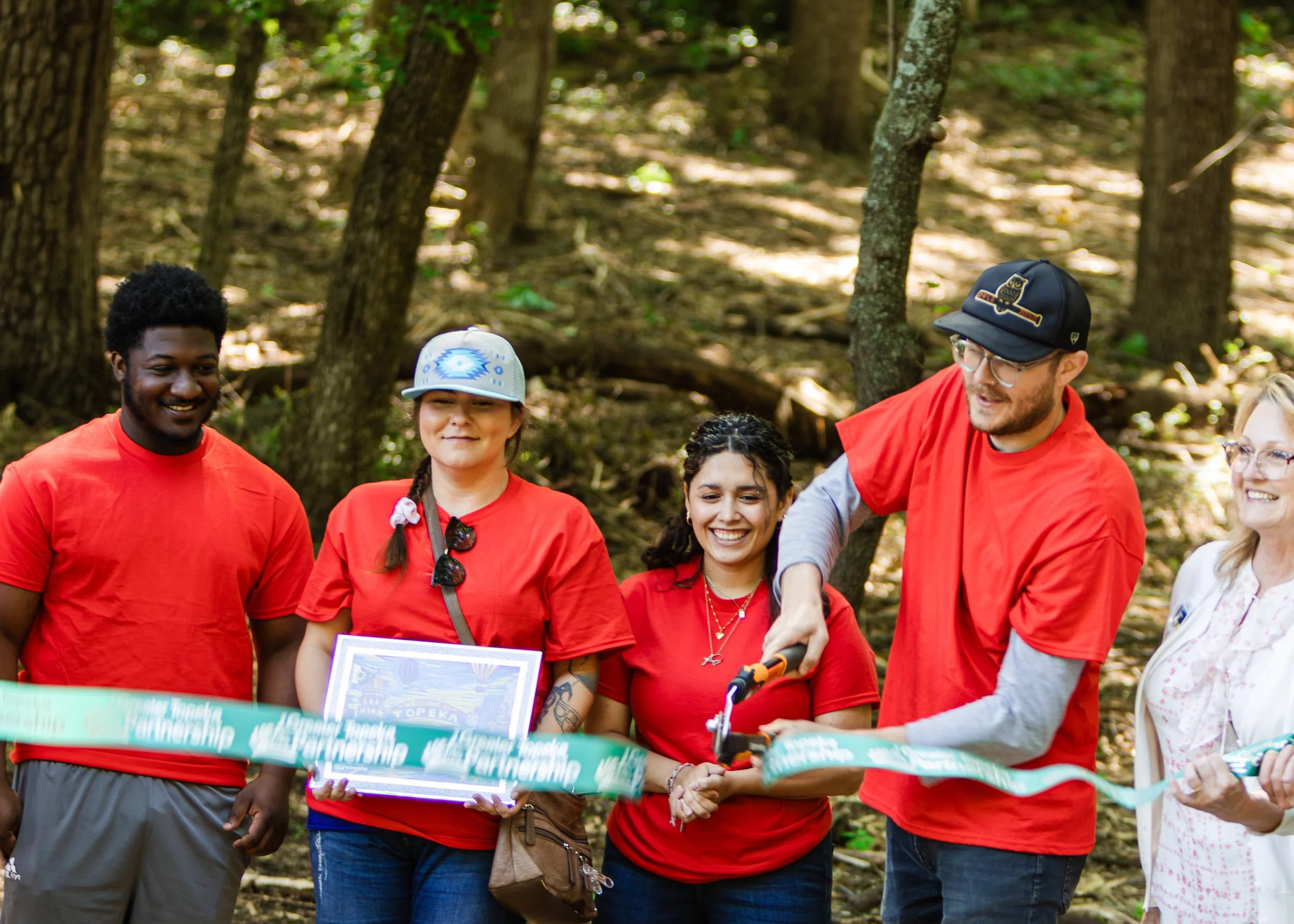 Group of people folding a ribbon at an outdoor event in a wooded area.