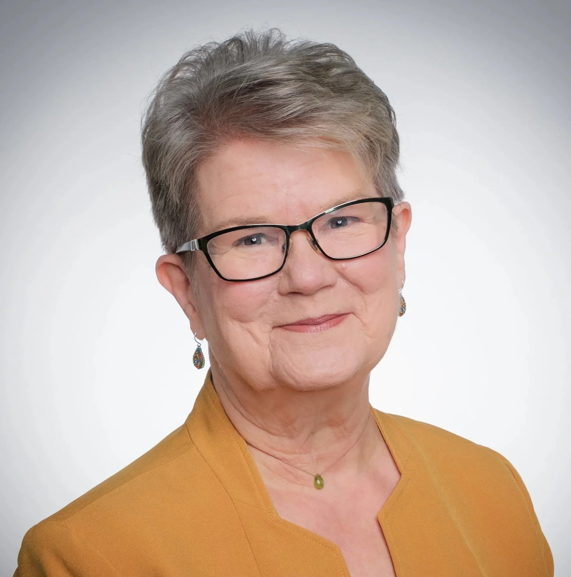 Close-up of a smiling older woman with short gray hair, wearing black-rimmed glasses, yellow blazer, matching jewelry, and a green necklace against a plain white background.