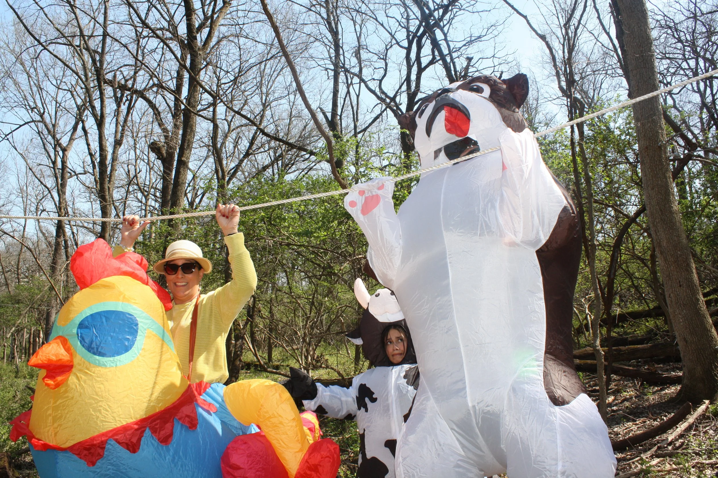 People celebrating outdoors in a park with colorful decorations, including a large cow-shaped lantern and a clown lantern, under leafless trees on a sunny day.