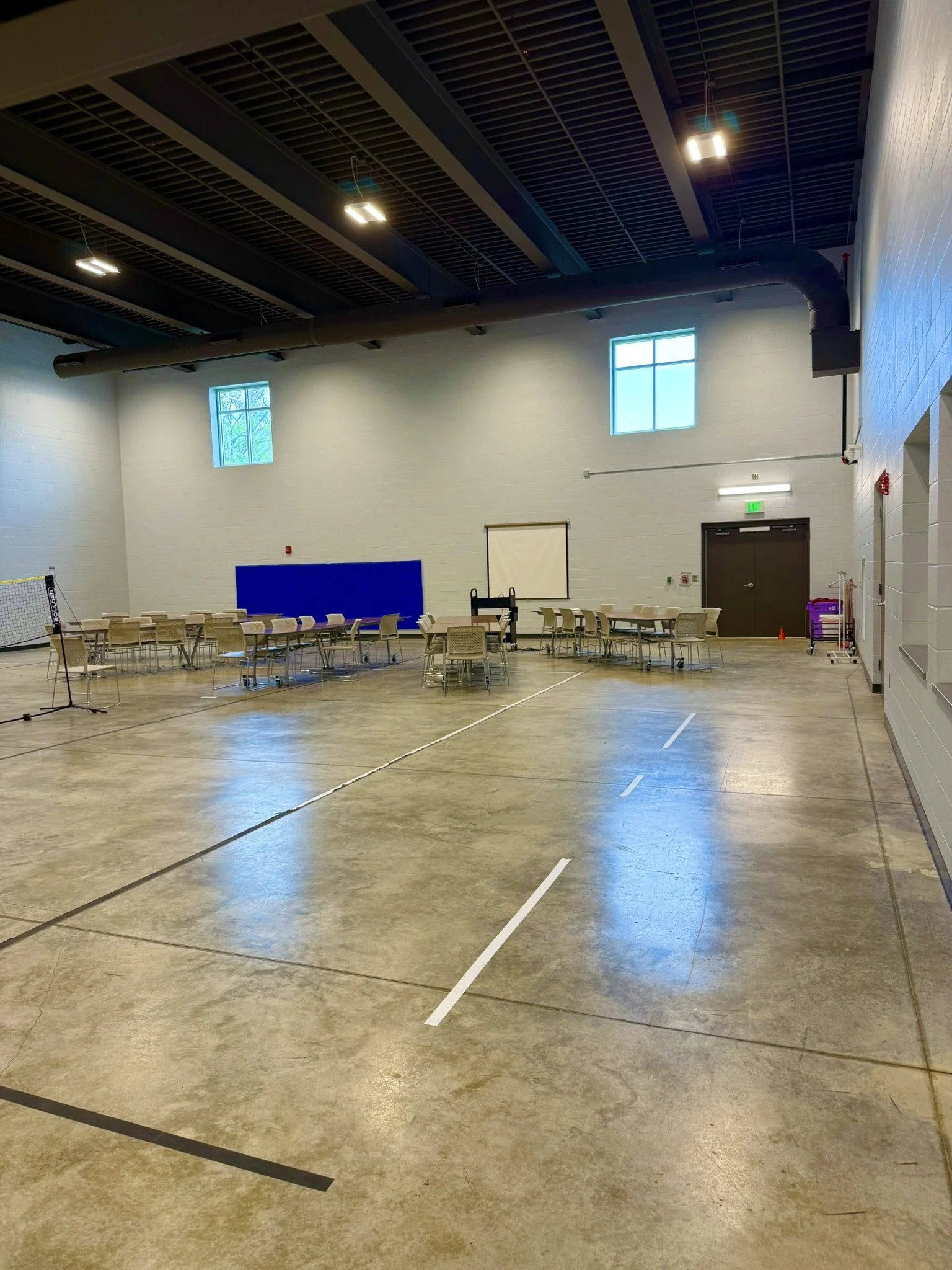 A large indoor gymnasium with high ceiling, two small windows, and tables and chairs arranged along the walls. The floor has markings for sports. The ceiling has exposed pipes and lighting fixtures.