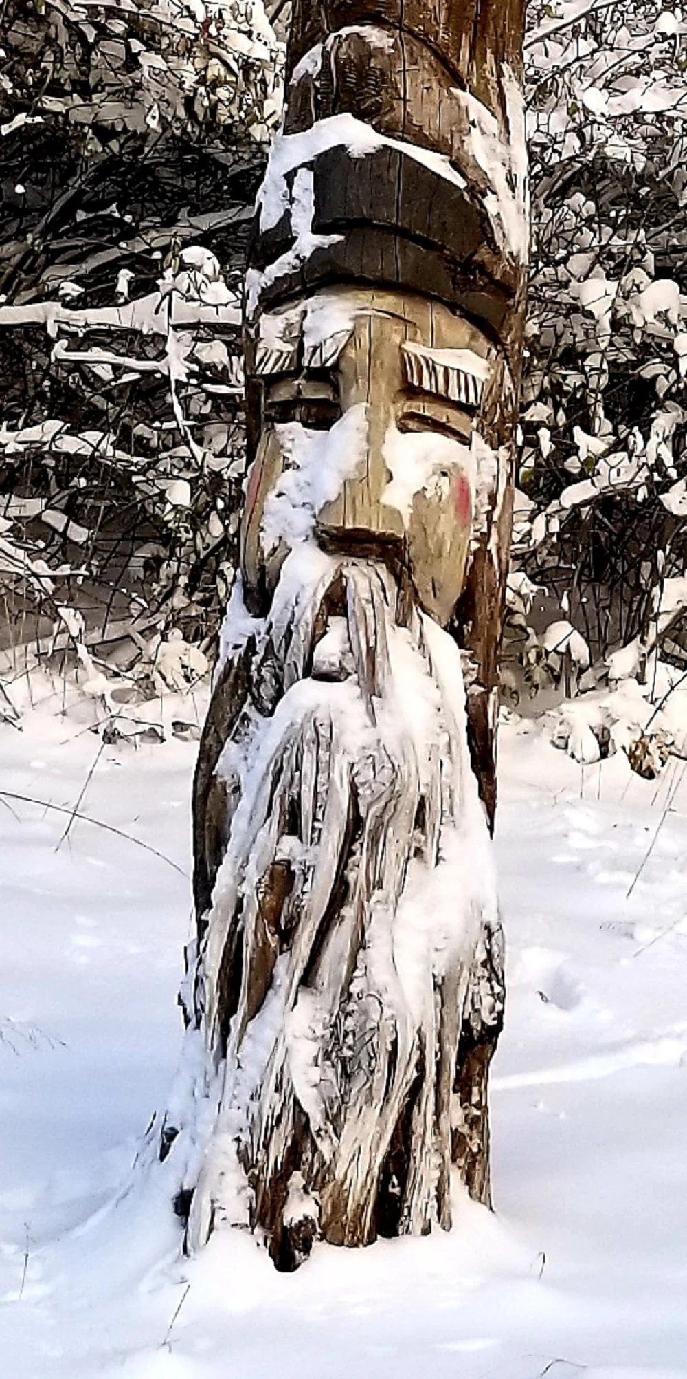 A carved wooden totem pole standing in the snow with snow-covered trees and bushes in the background.