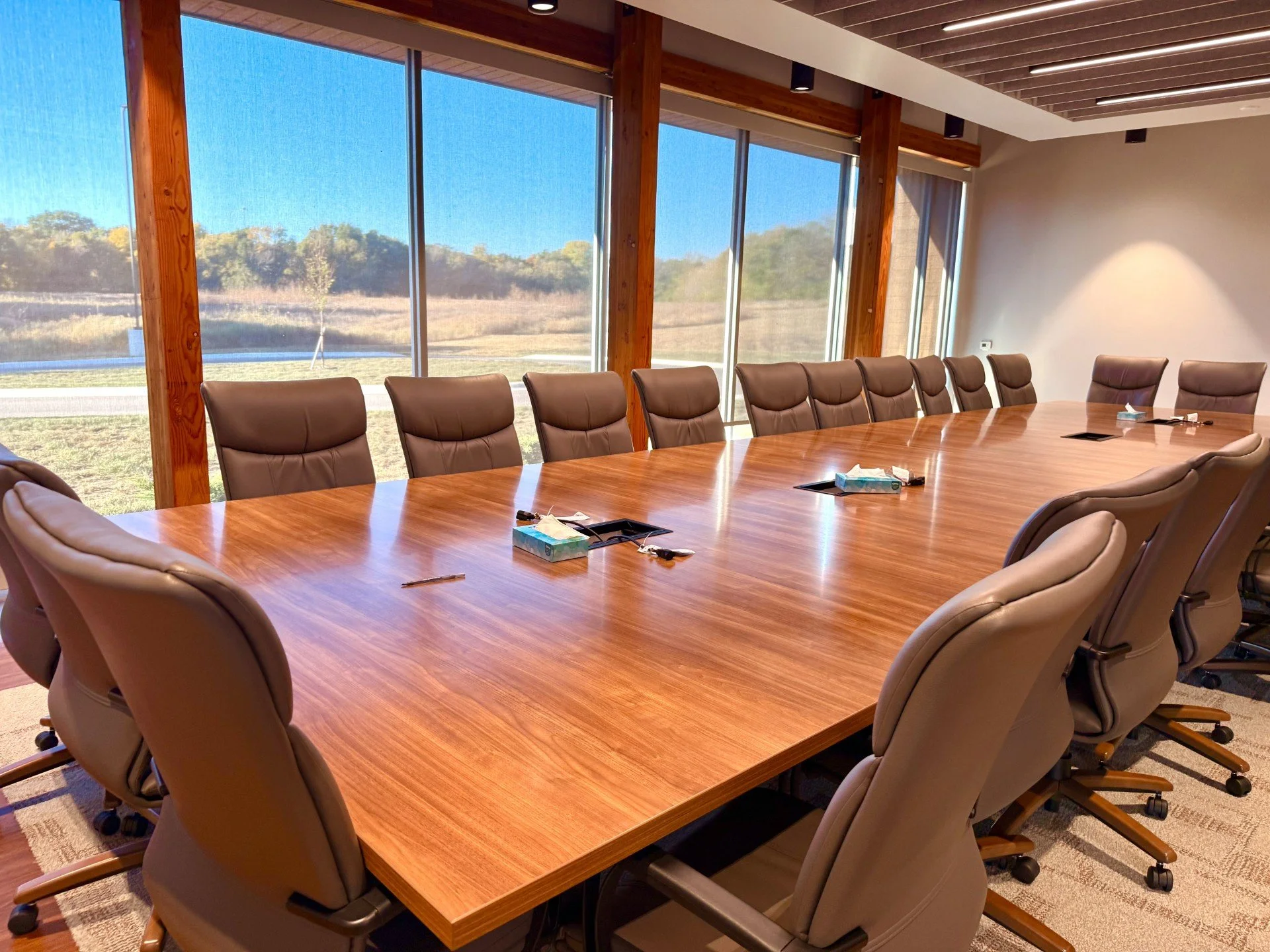 Conference room with a large wooden table and beige leather chairs, large windows showing an outdoor landscape with trees, tissue boxes and writing instruments on the table.