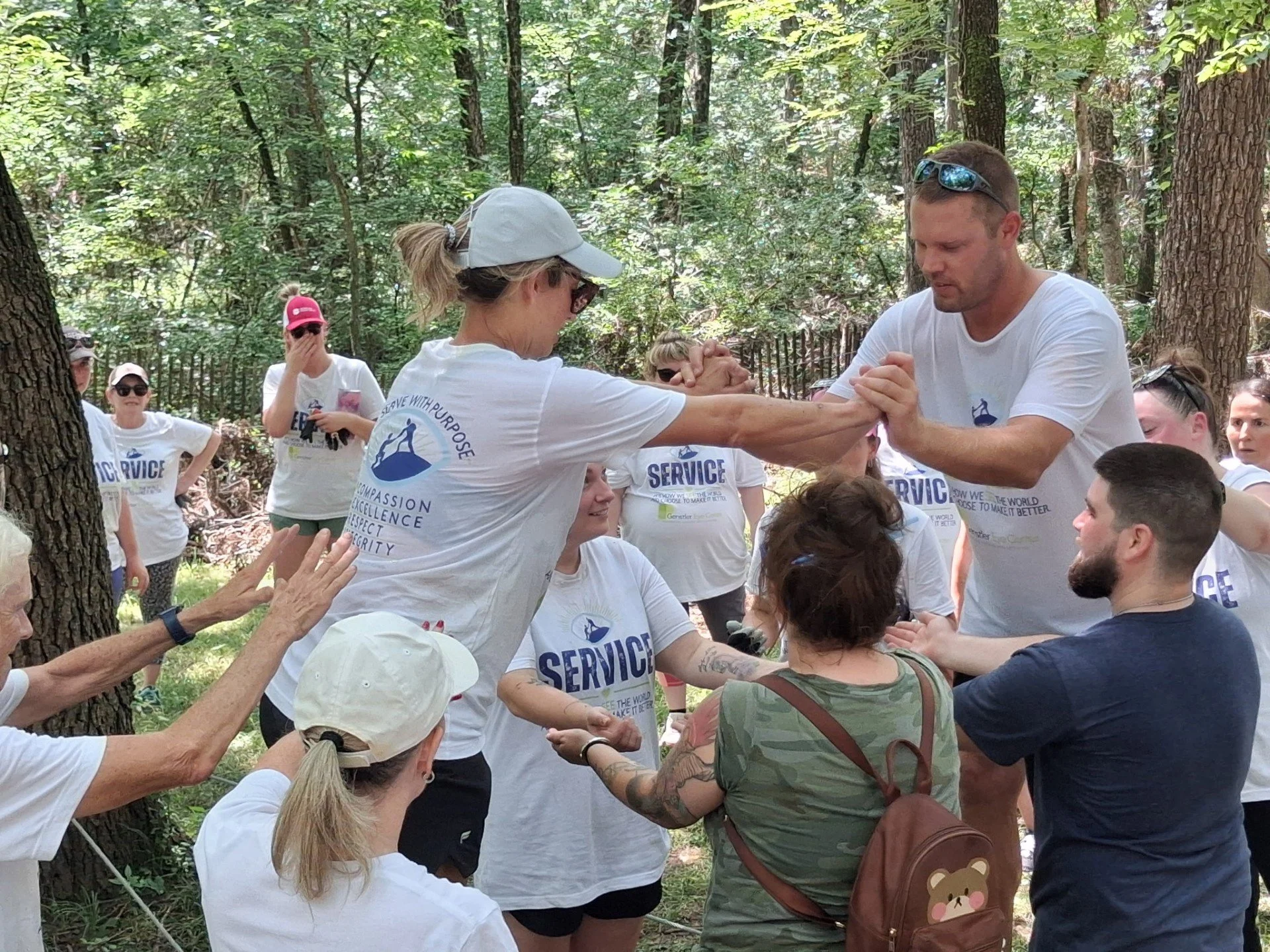 A group of people outdoors in a forest, participating in an event wearing white t-shirts with the words 'SERVICE' and other text, some wearing hats and sunglasses. Two people are engaged in arm wrestling.