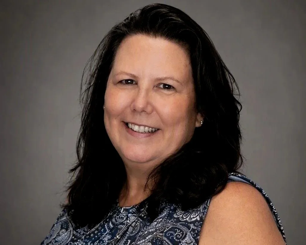 Portrait of a smiling woman with dark wavy hair wearing a blue patterned sleeveless top against a plain gray background.