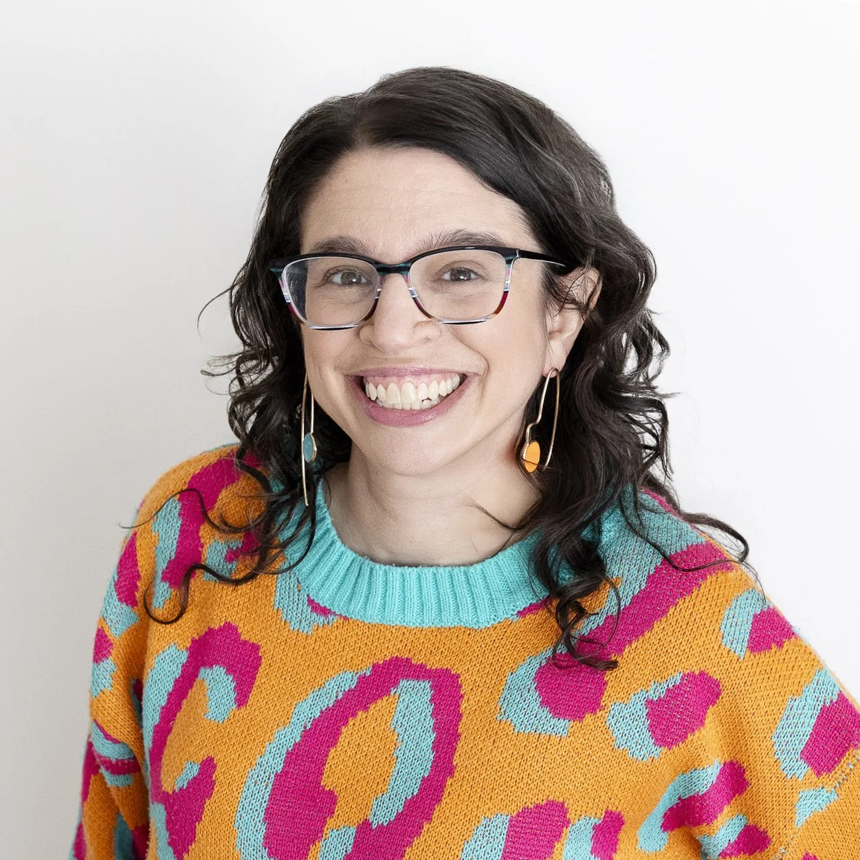 A woman with dark, curly hair wearing glasses, colorful earrings, and a multicolored sweater with a bright smile.