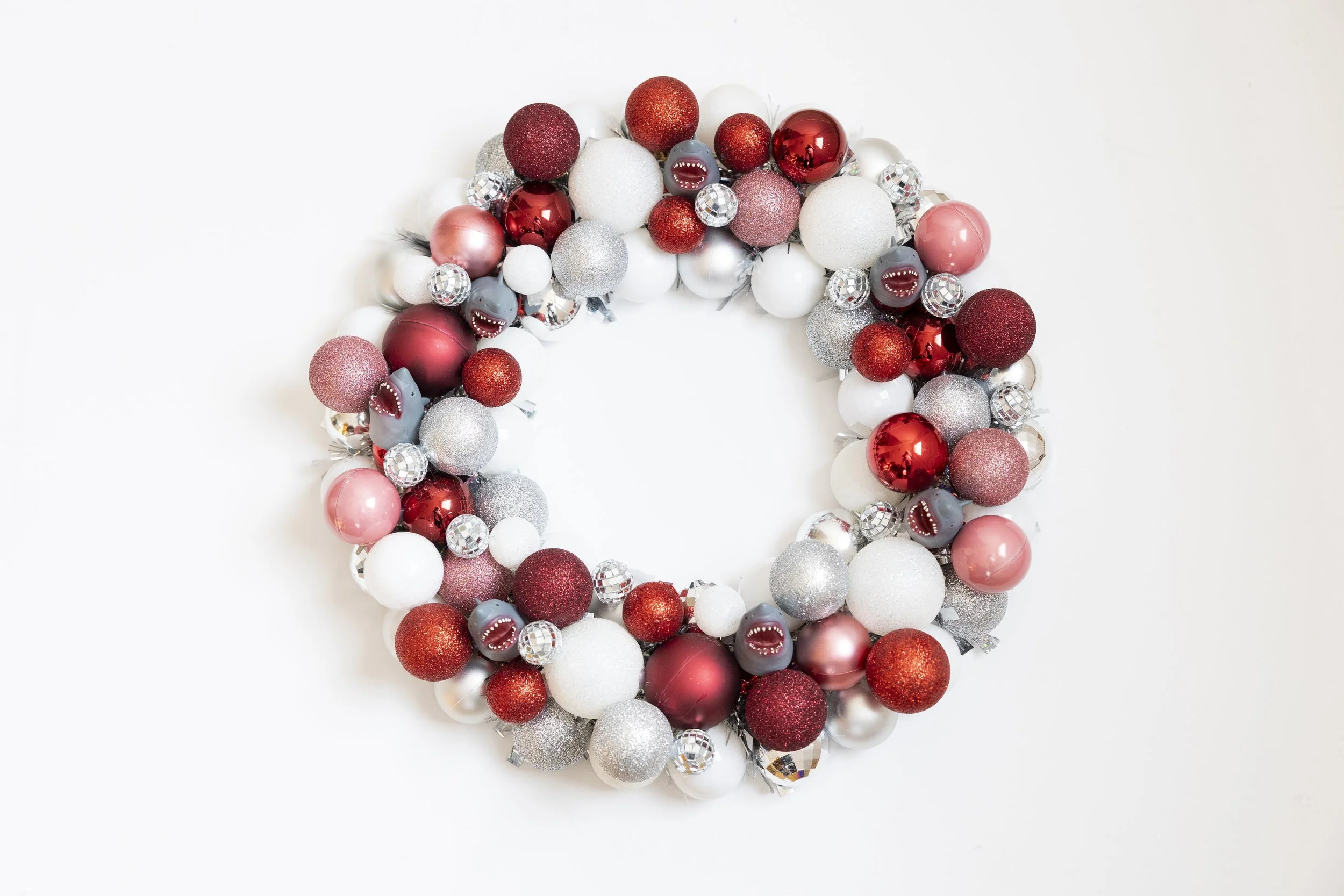 Holiday shark wreath made of red, white, silver, pink, and disco ball Christmas ornaments on a white background.