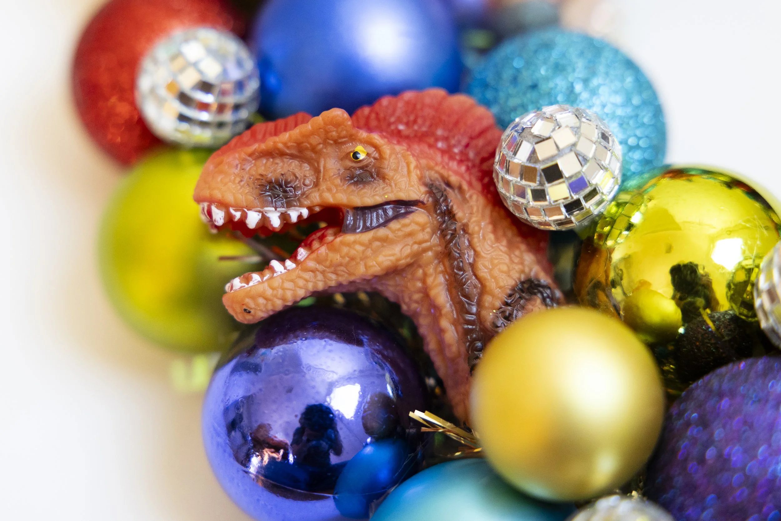 Close-up of various colorful ornaments on a wreath including a small dinosaur figurine, metallic and glittery disco balls.