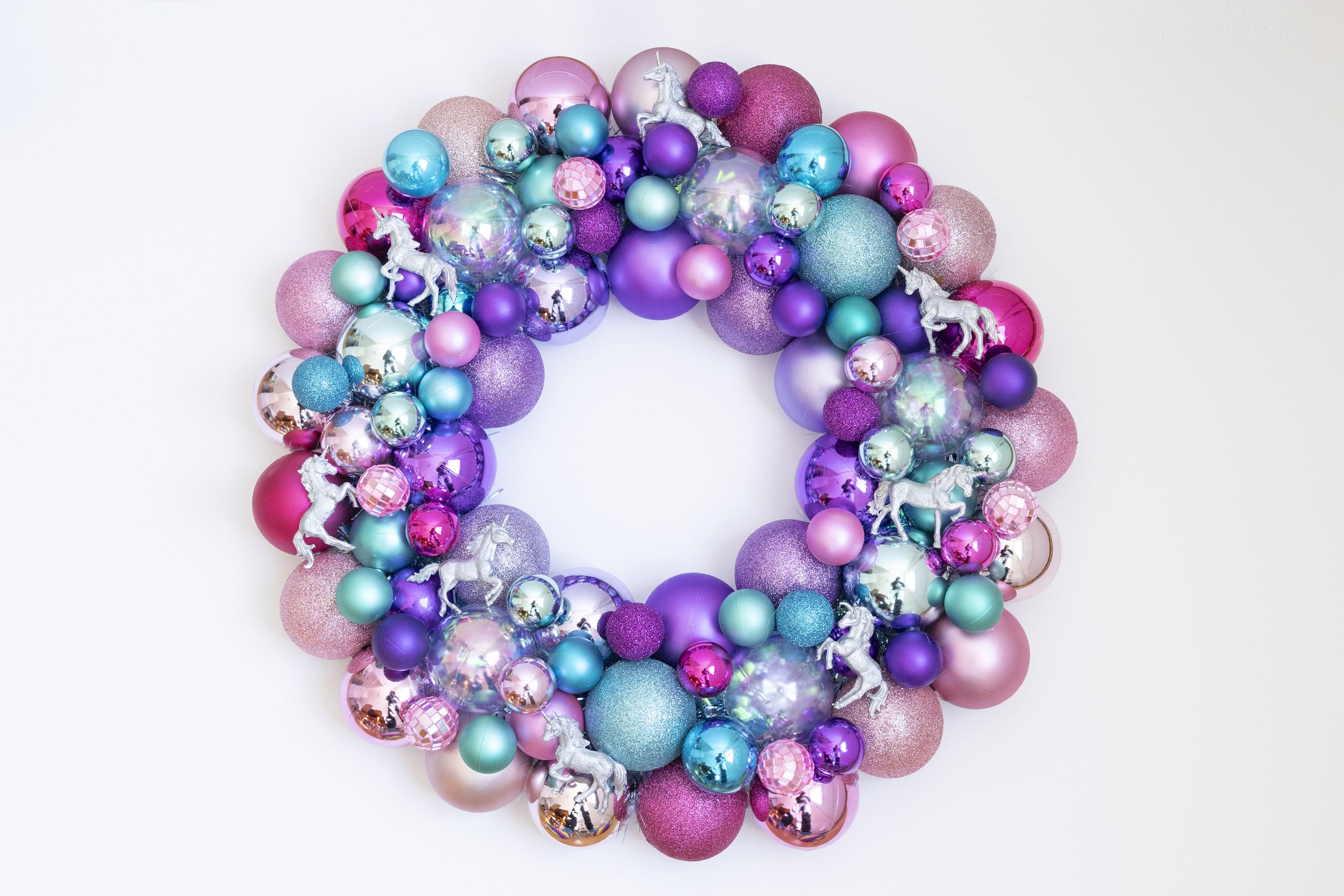 Colorful decorative wreath made of pink, purple, turquoise, and glittery ornaments, including unicorn figurines, set against a white background.