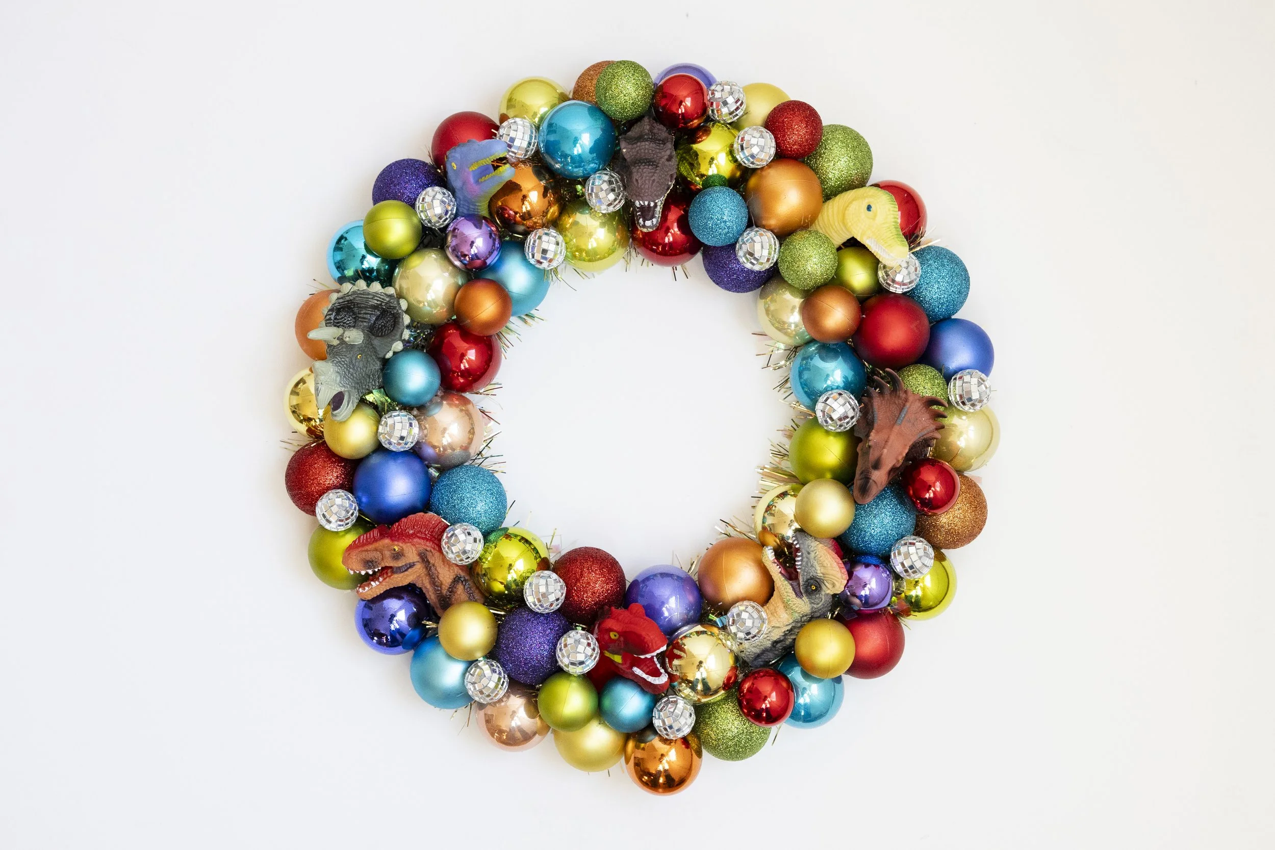 Colorful dinosaur-themed Christmas wreath made of various sized shiny and glittery ornaments with dinosaur figures, against a white background.