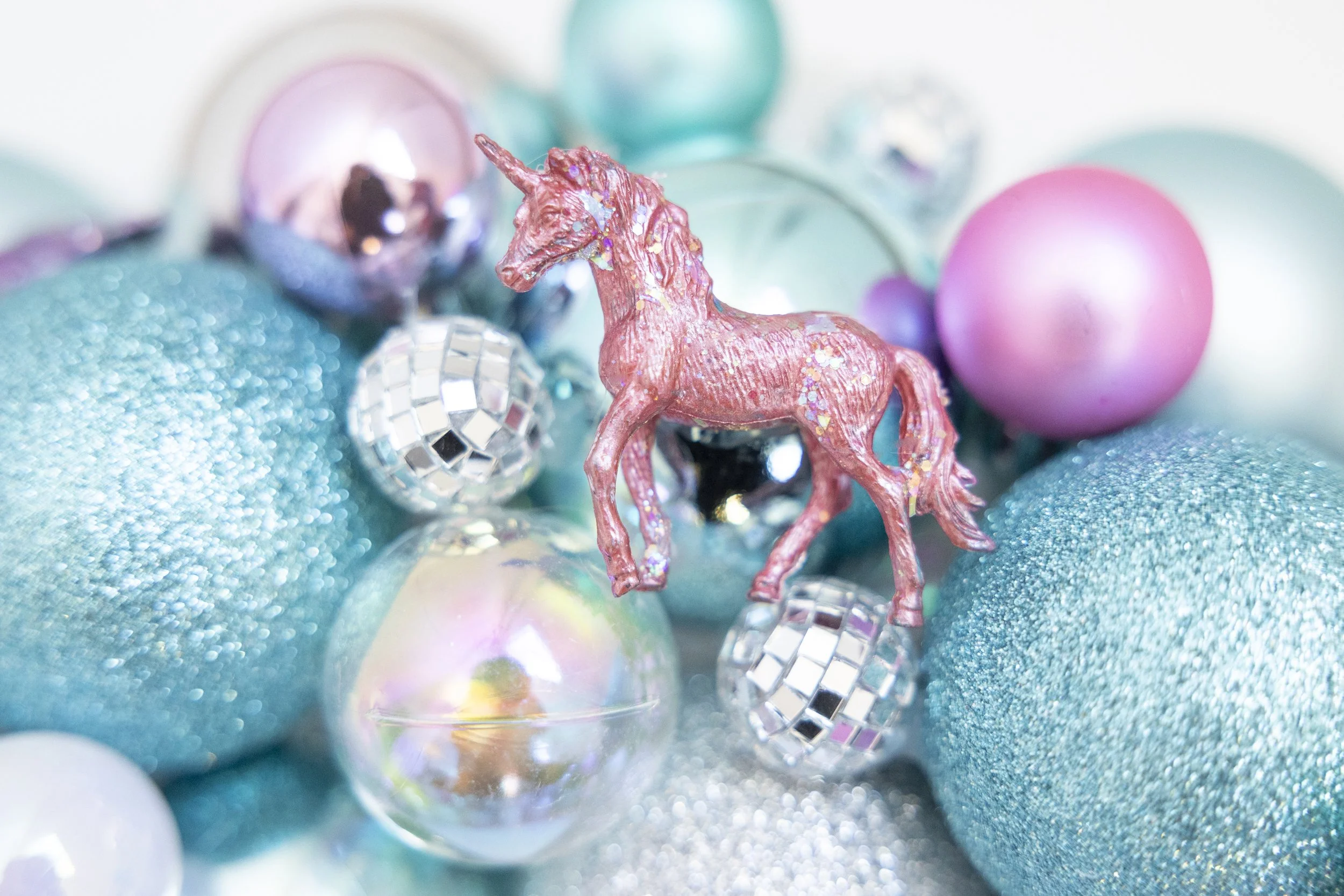 Close-up of a pink unicorn figurine with glitter, surrounded by colorful decorative ornaments and shiny disco balls against a soft, blurred background.