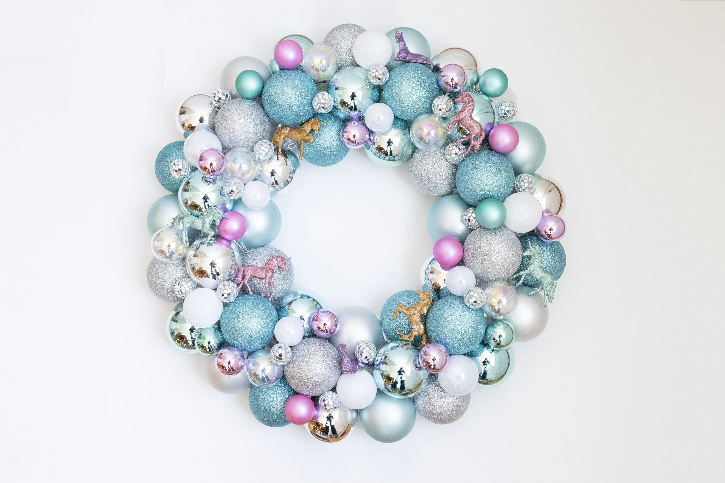 A decorative wreath made of blue, silver, pink, and iridescent ornaments and small unicorn figures against a white background.