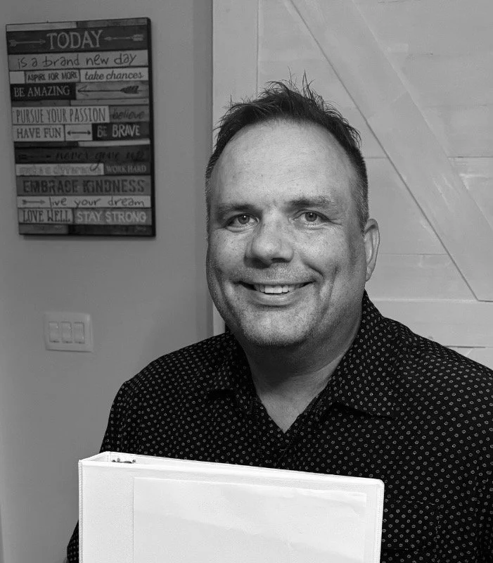A smiling man with short hair and light skin, wearing a dark, polka-dot shirt, holding a white folder, standing indoors near a wall with a motivational quote board in the background.