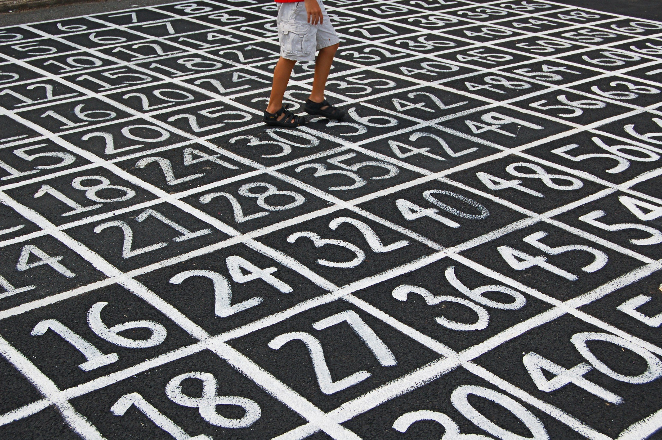 A child standing on a black asphalt court with white numbered lines and grids, possibly a hopscotch game or similar drawing.