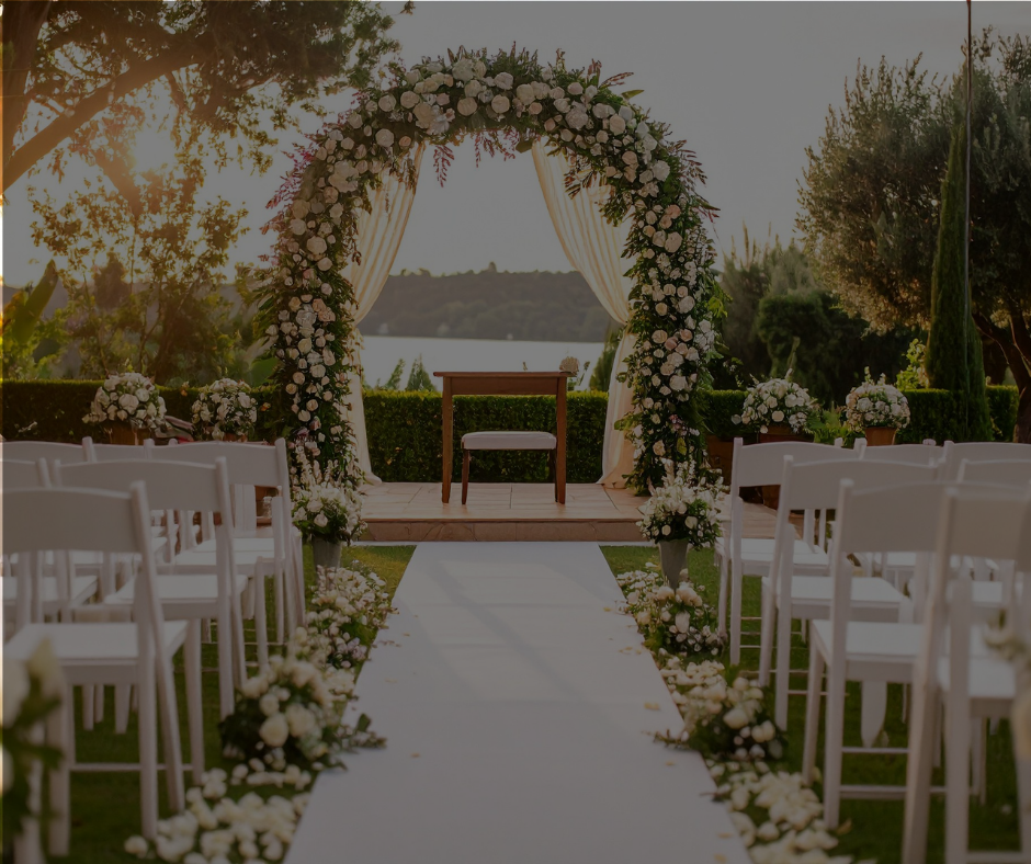 Outdoor wedding ceremony setup with white chairs, floral arch, and aisle with flower arrangements, overlooking a body of water at sunset.
