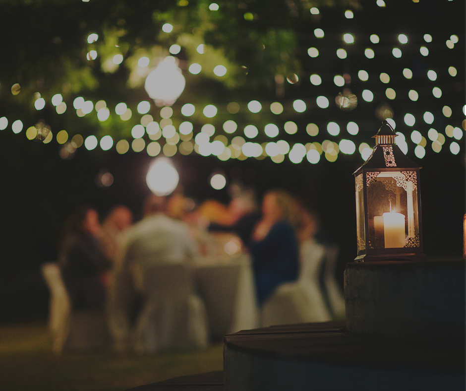 A lit candle inside a decorative lantern on a table at an outdoor evening gathering with blurred guests and string lights in the background.