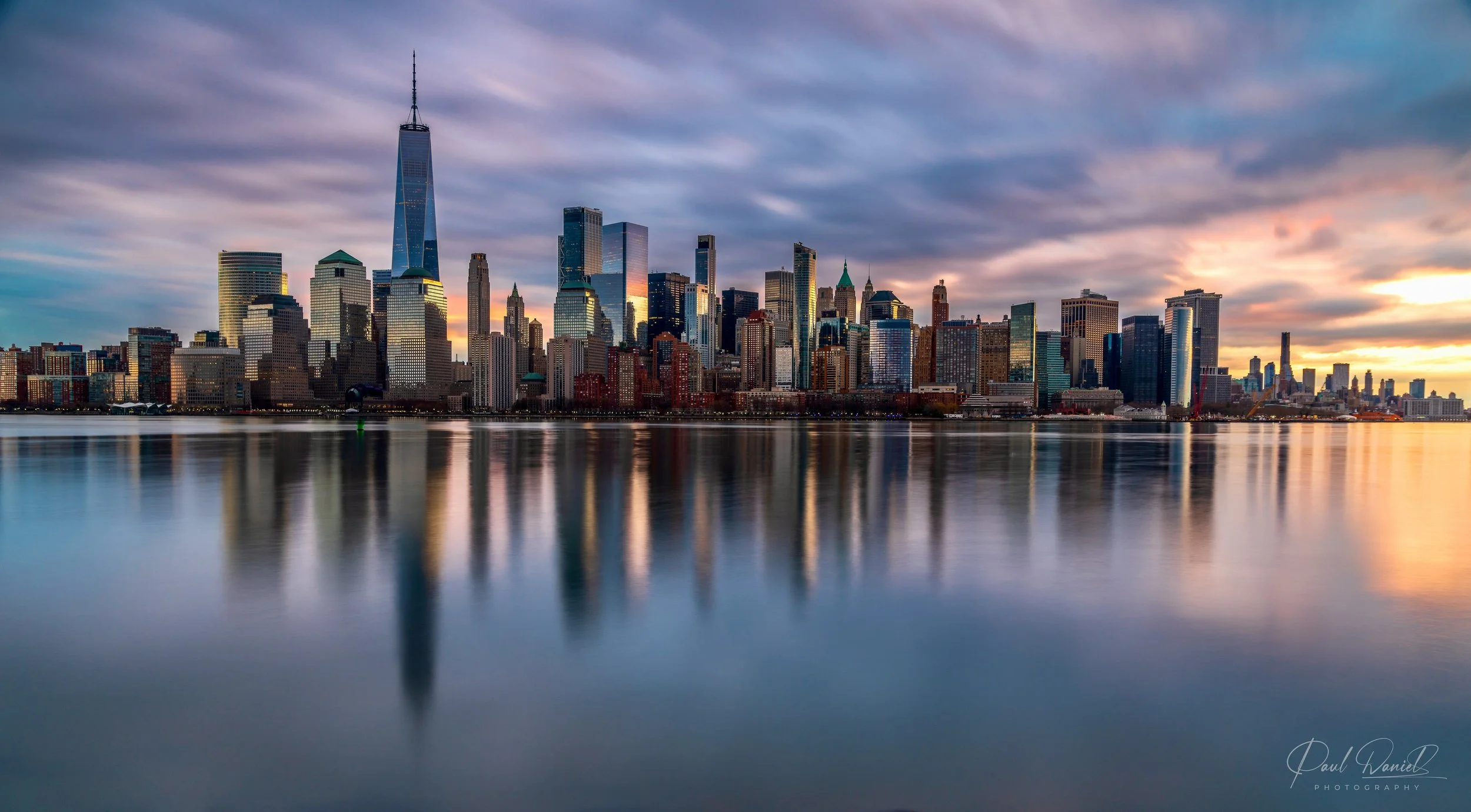 Panoramic sunset landscape photography of the Lower Manhattan skyline and One World Trade Center reflected in the Hudson River as seen from the New Jersey waterfront. 