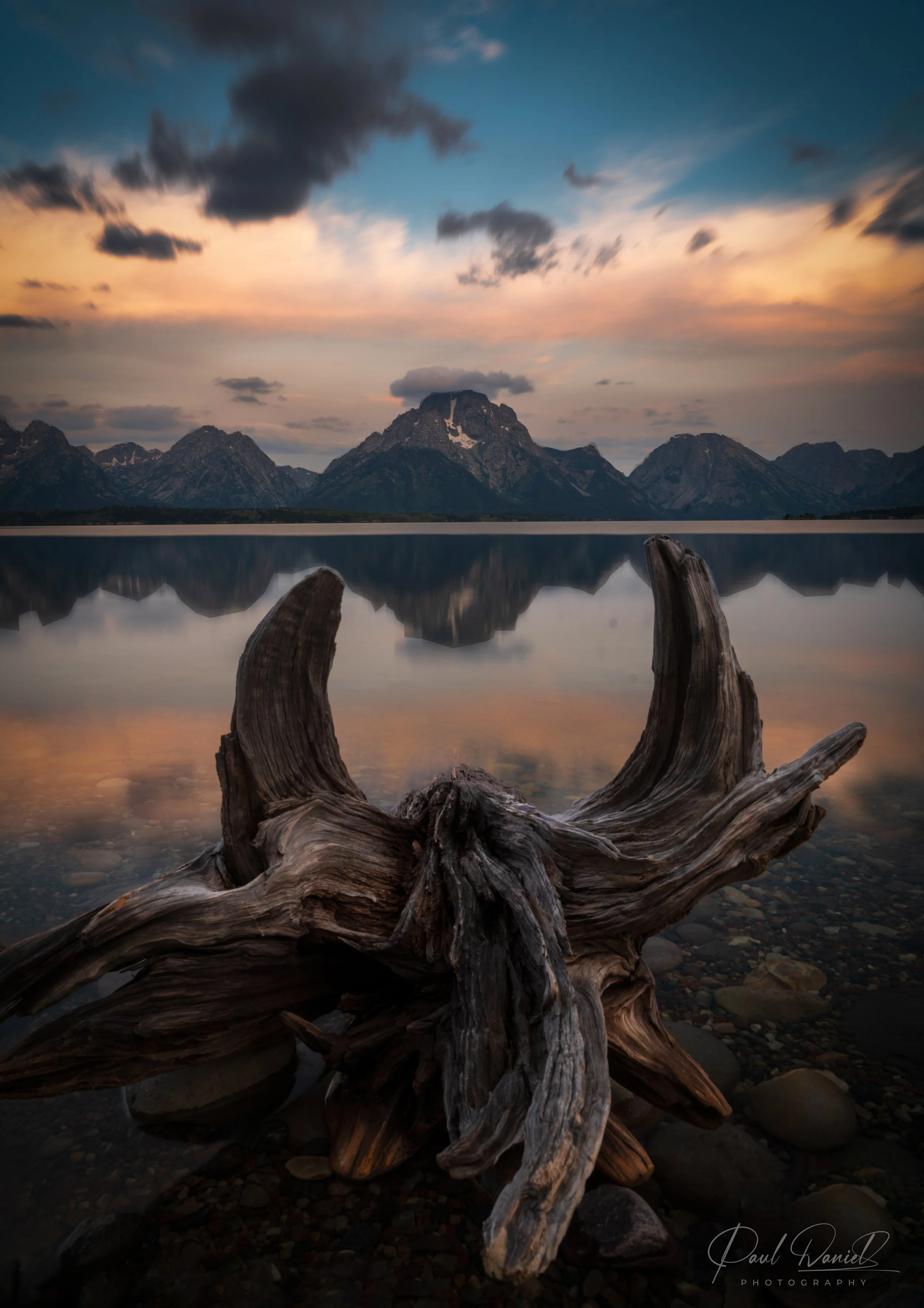 Vertical fine art landscape photograph of the Teton Range reflected in calm lake waters framed by weathered driftwood at sunset in Grand Teton National Park, Wyoming (USA)