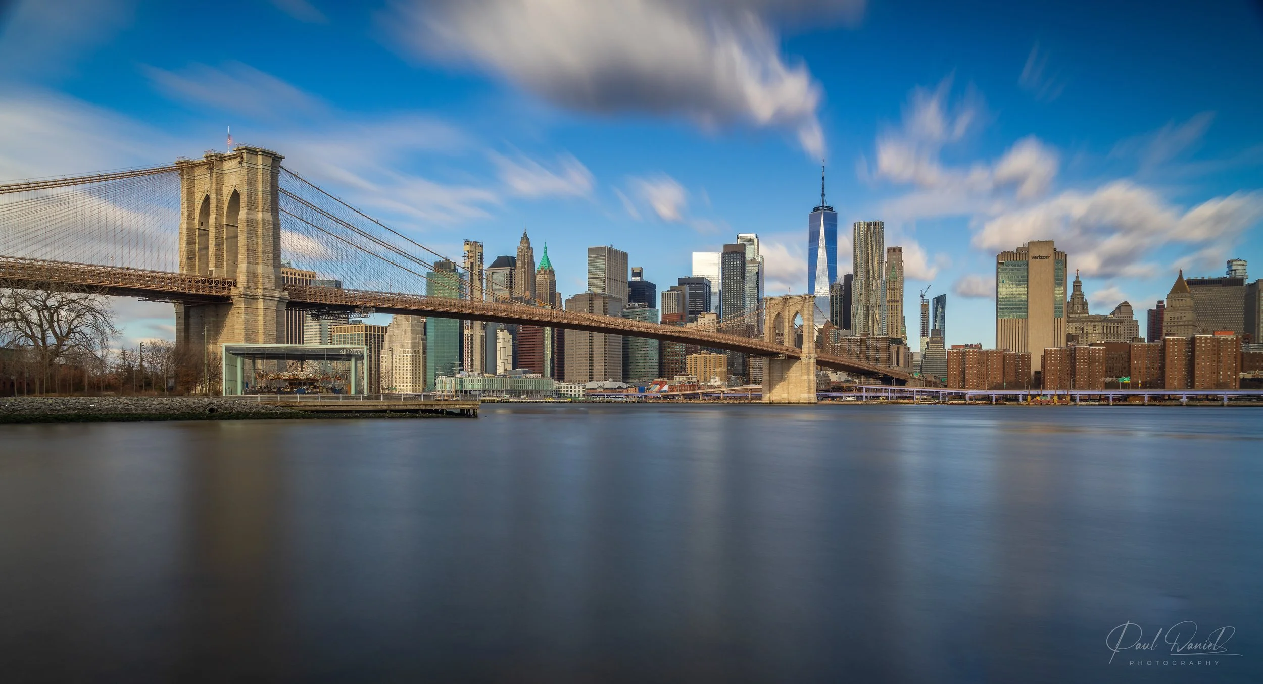 Panoramic daytime view of the Brooklyn Bridge spanning the East River toward the Lower Manhattan skyline and One World Trade Center, as seen from Brooklyn Bridge Park. 