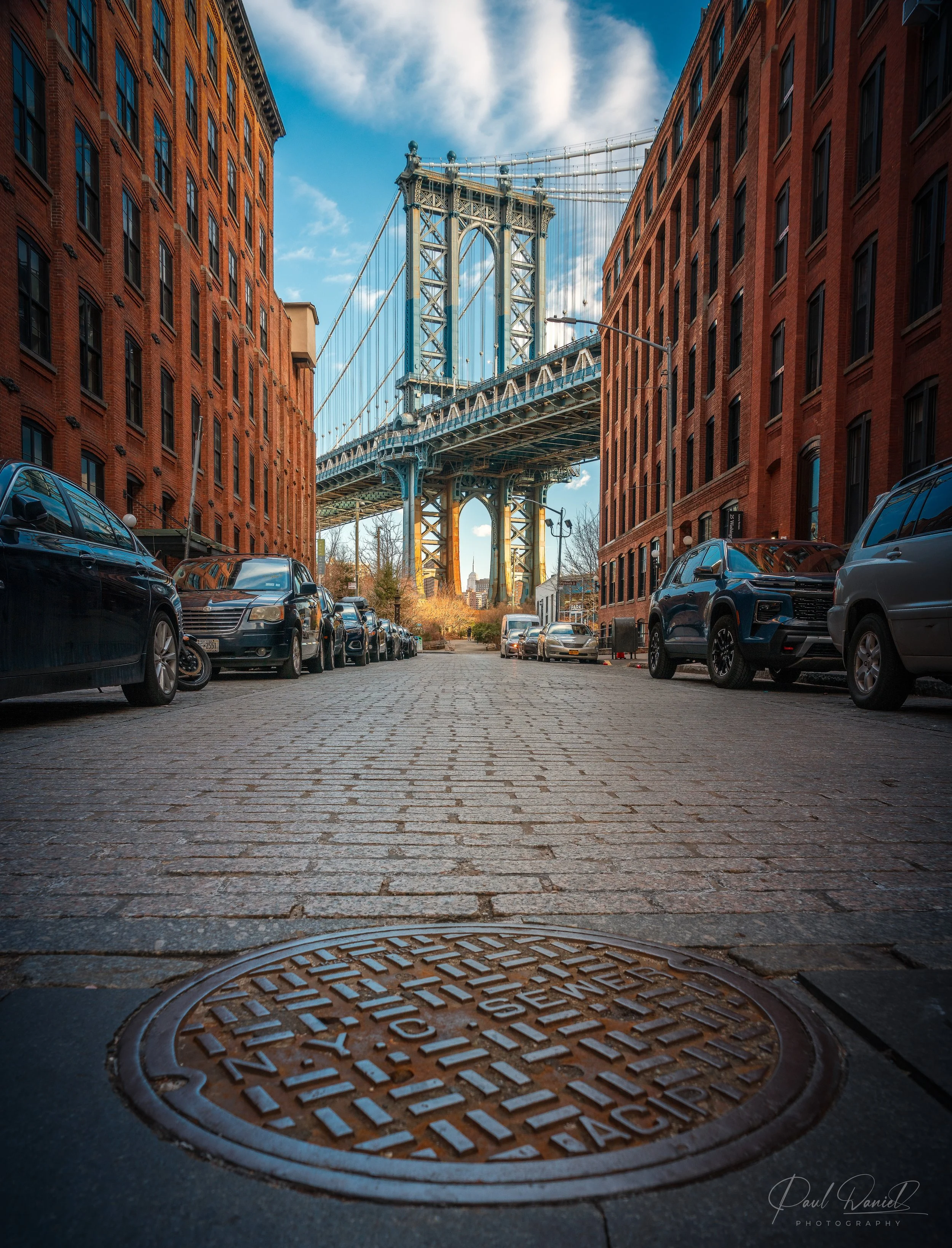Ground-level, wide-angle street photography of Washington Street in DUMBO, Brooklyn, framing the Manhattan Bridge and cobblestone street with a manhole cover in the foreground. 