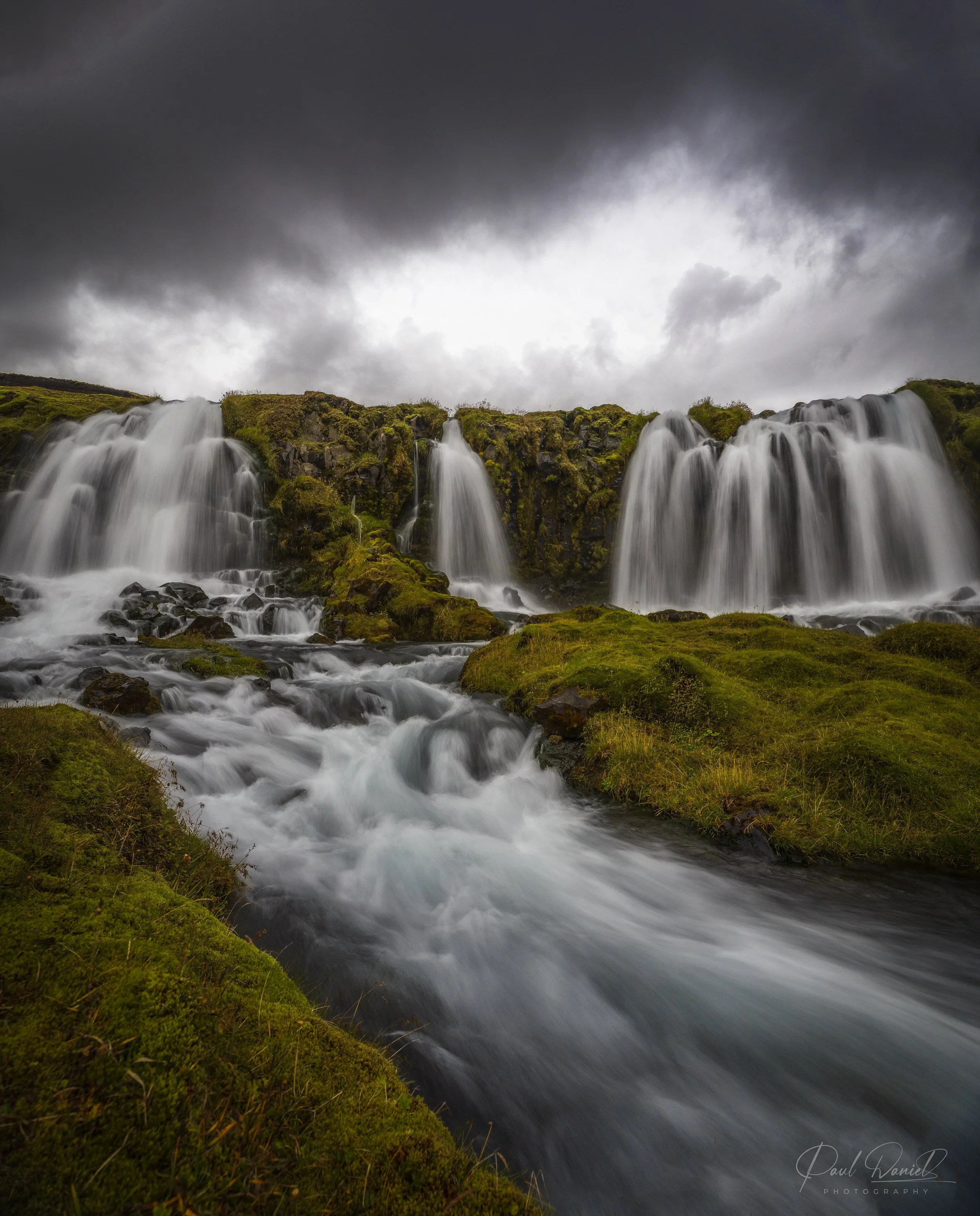Dramatic long-exposure landscape photography of a cascading waterfall flowing over moss-covered volcanic rock under stormy overcast skies in Iceland.