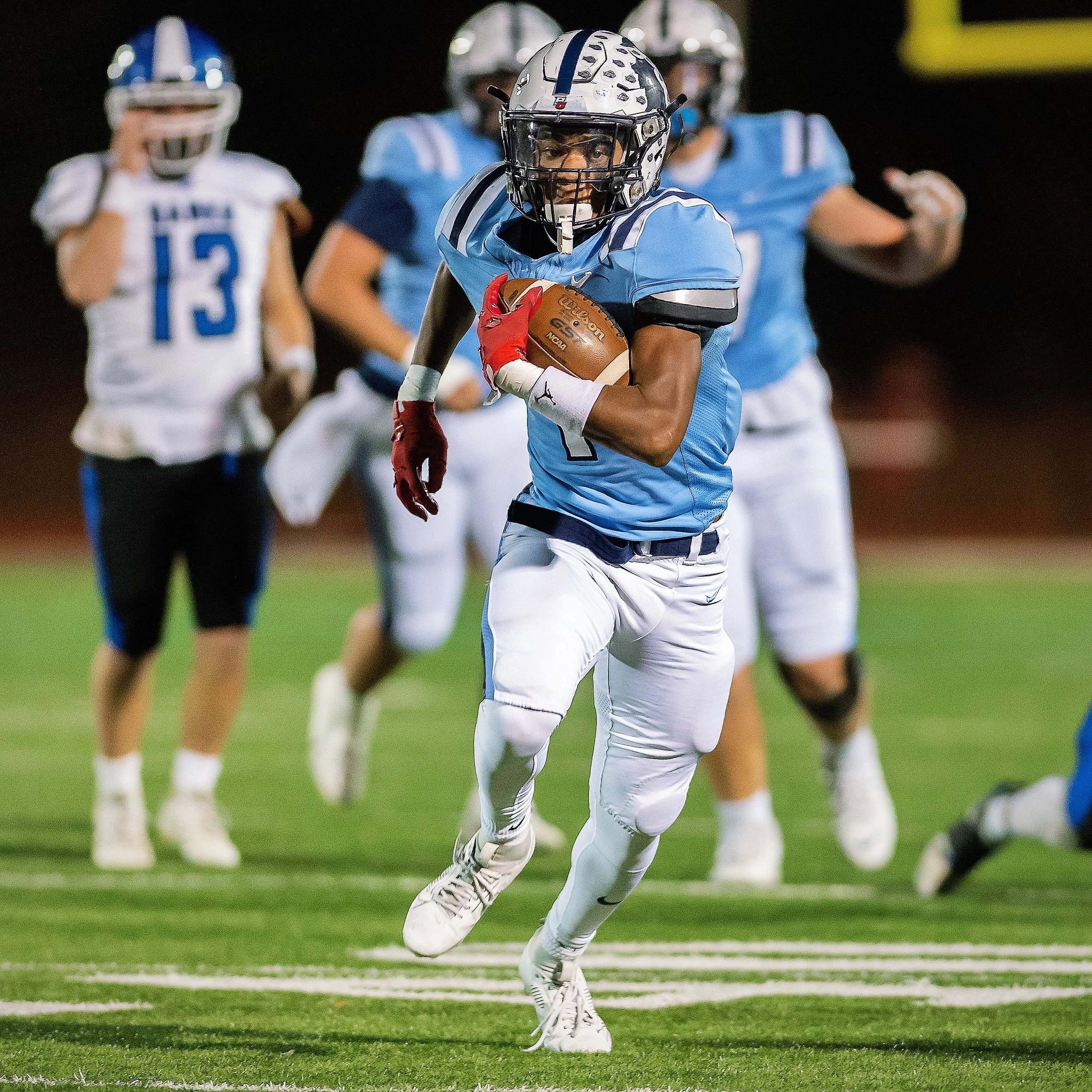 High school football action shot featuring a running back breaking through coverage down the field under Friday night stadium lights. 