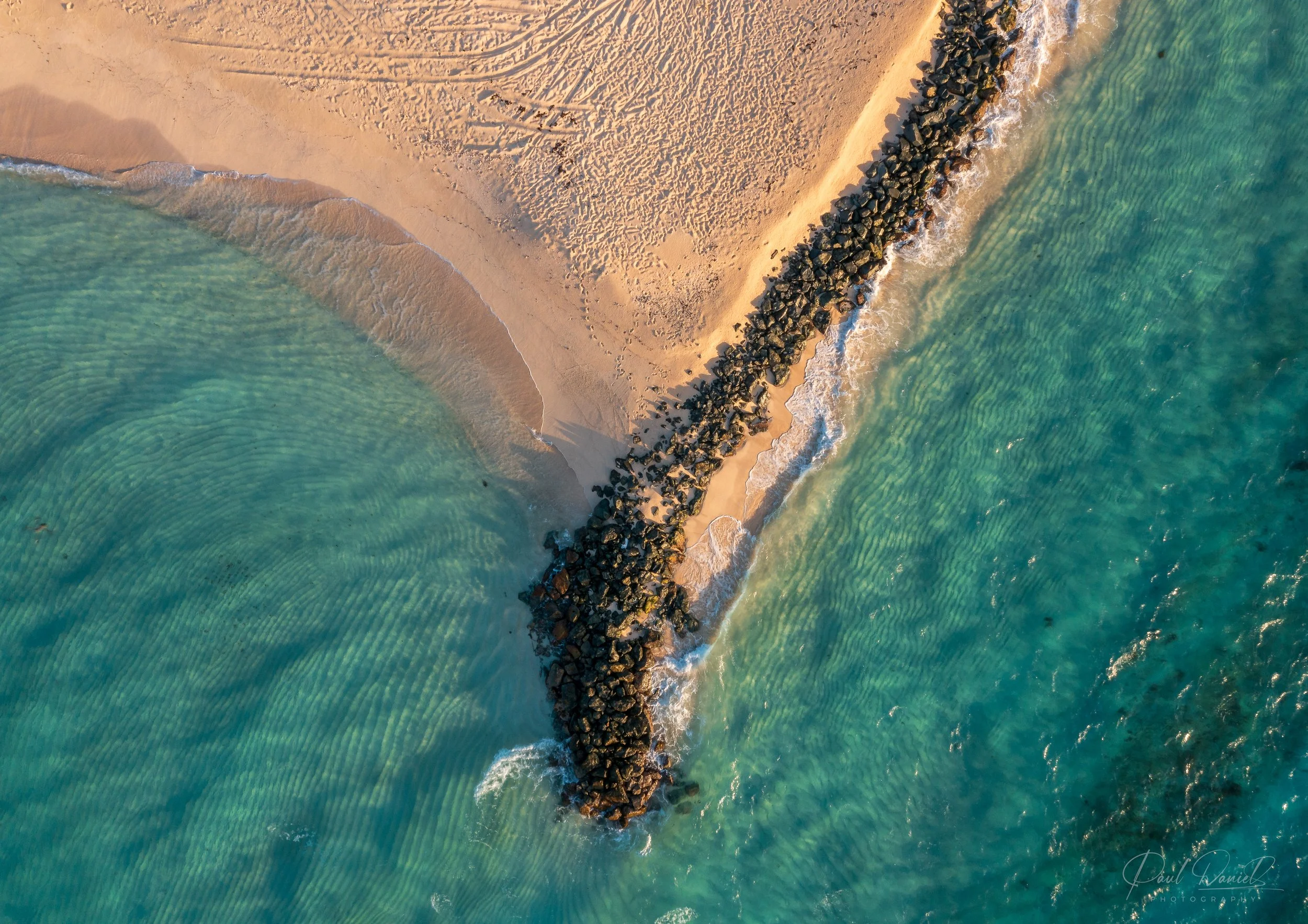 Top-down aerial drone photograph of a private sandy beach, rock jetty, and clear-crystal turquoise waters on Necker Island in the British Virgin Islands. 