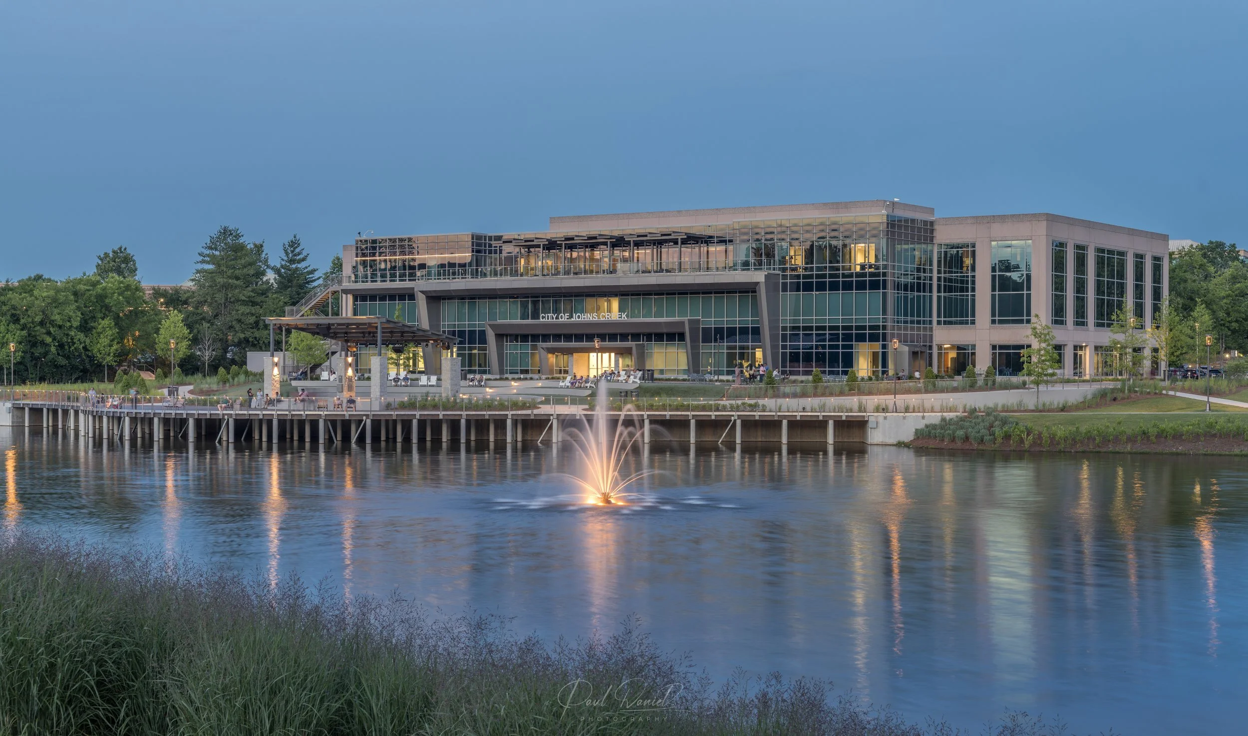 Twilight landscape photography of Johns Creek City Hall, featuring the illuminated lake, fountain, boardwalk deck, and modern municipal building in Johns Creek, GA