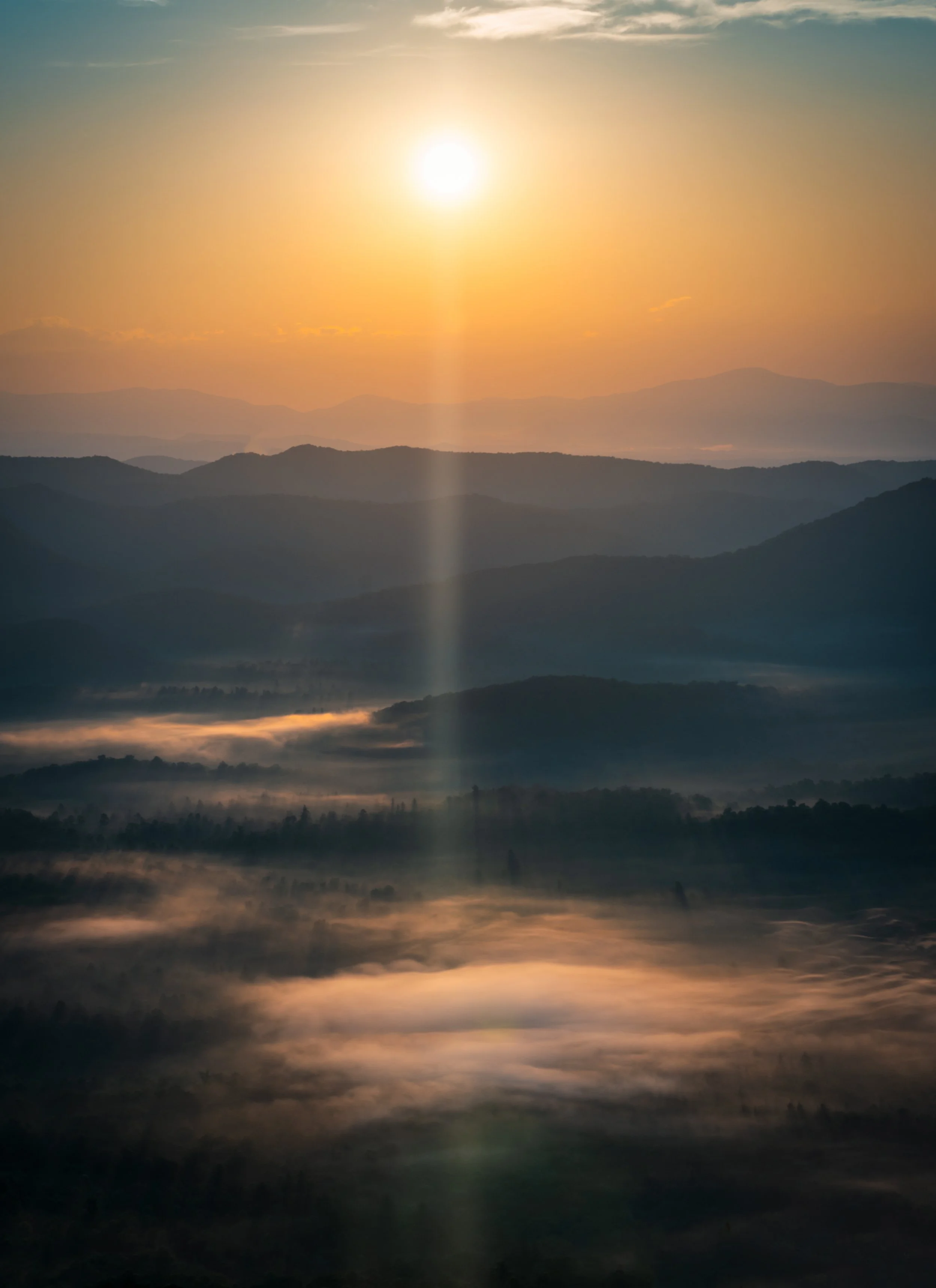 Vertical morning landscape photograph of low valley fog and mountain ridge layers glowing at sunrise along the Blue Ridge Parkway. 