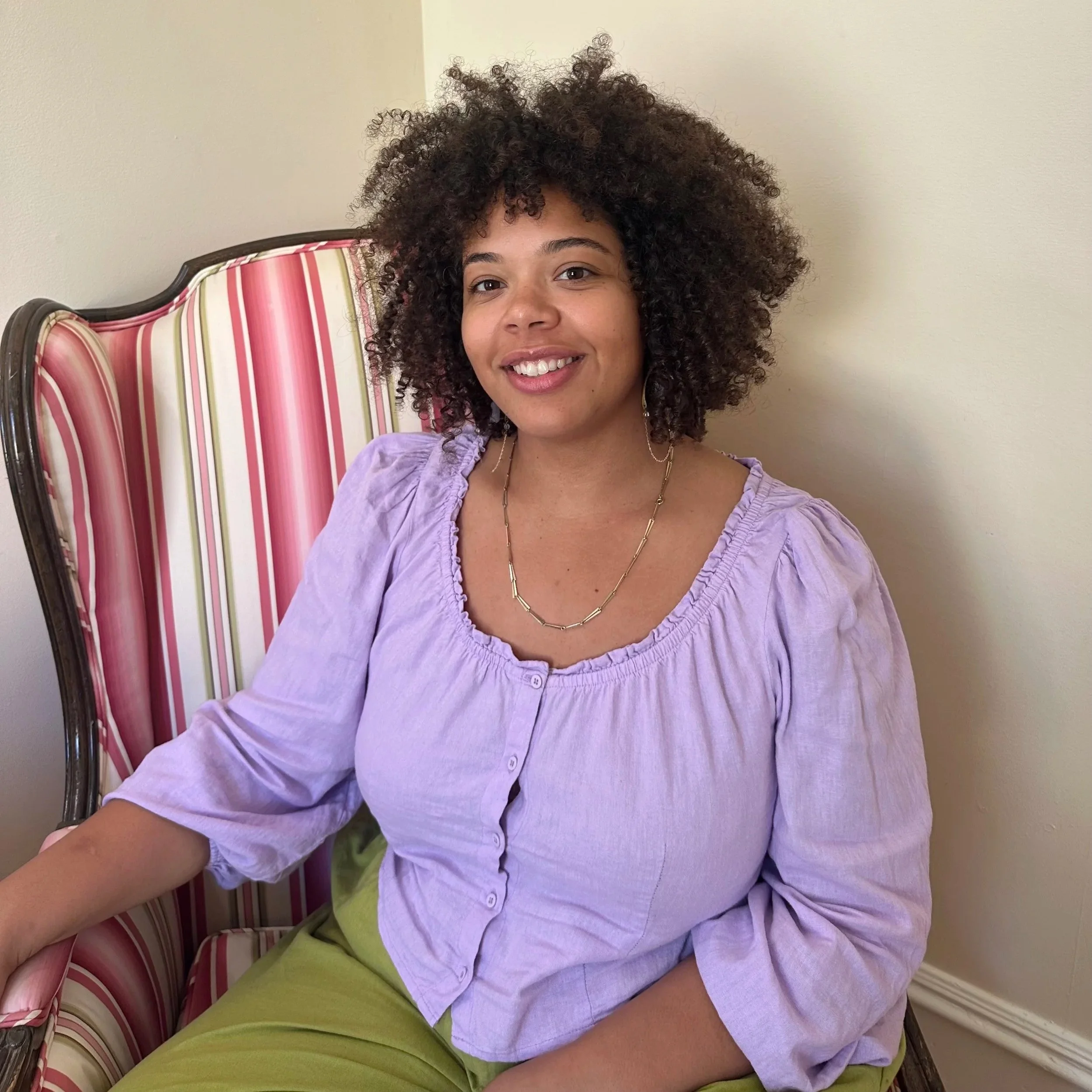 Greer Sargeant, a multiracial woman with curly hair sitting on a striped pink and green chair, smiling, wearing a lilac blouse and lime green pants.