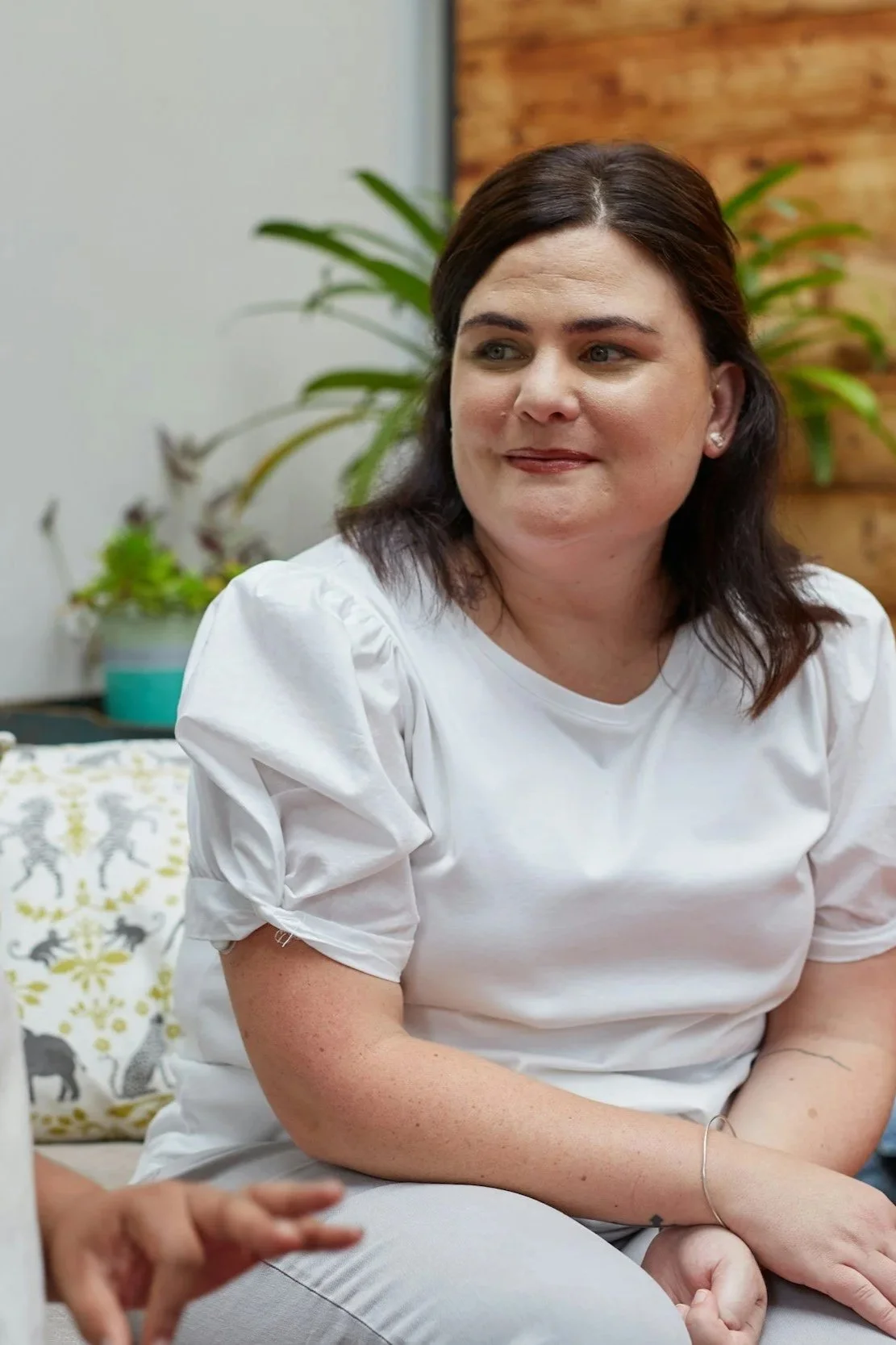 A woman with dark hair sitting indoors, wearing a white top with puffed sleeves, with green plants and a wooden wall in the background.