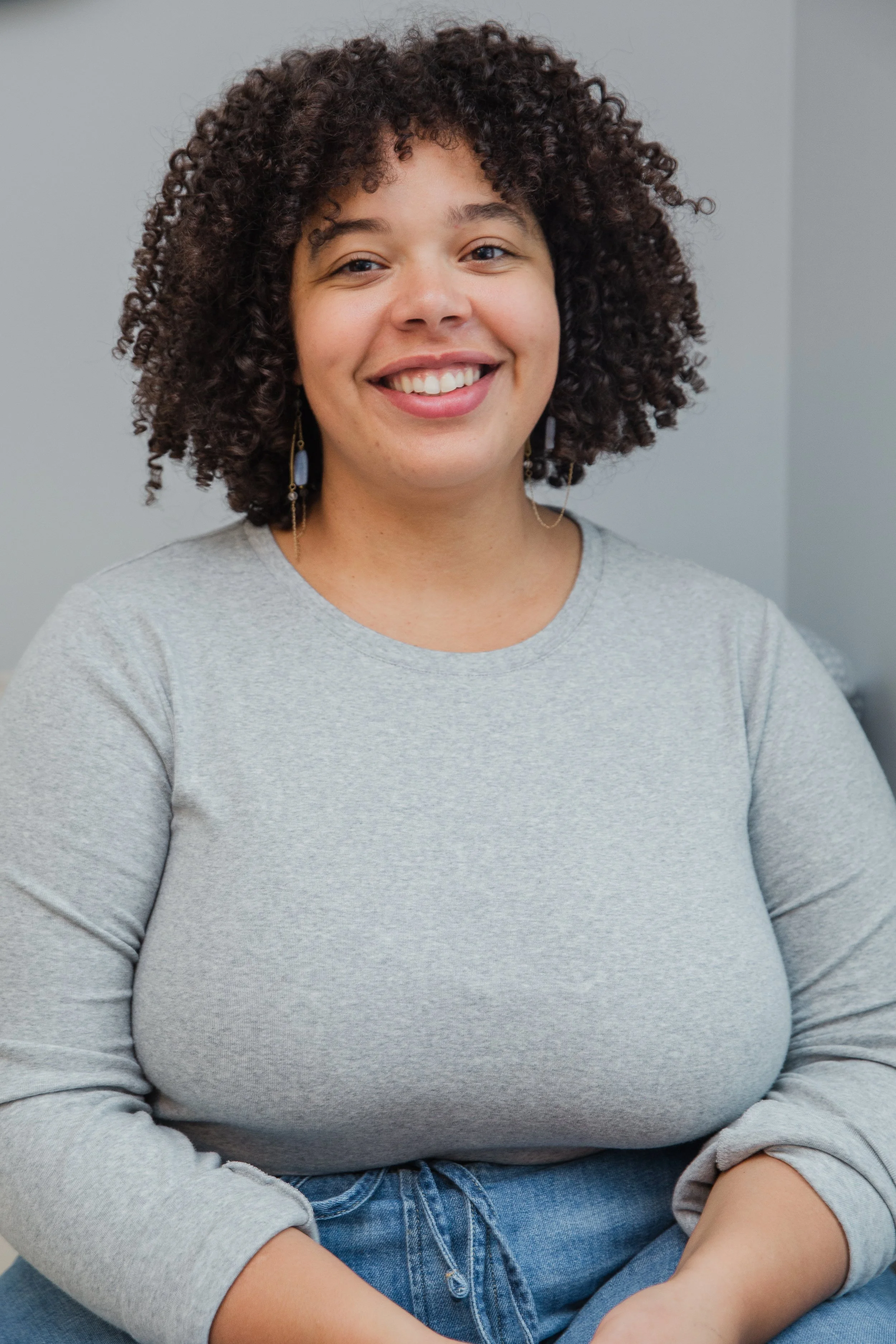 Greer Sargeant, a woman with curly hair smiling, wearing a gray long-sleeve shirt and blue jeans, against a plain gray background.