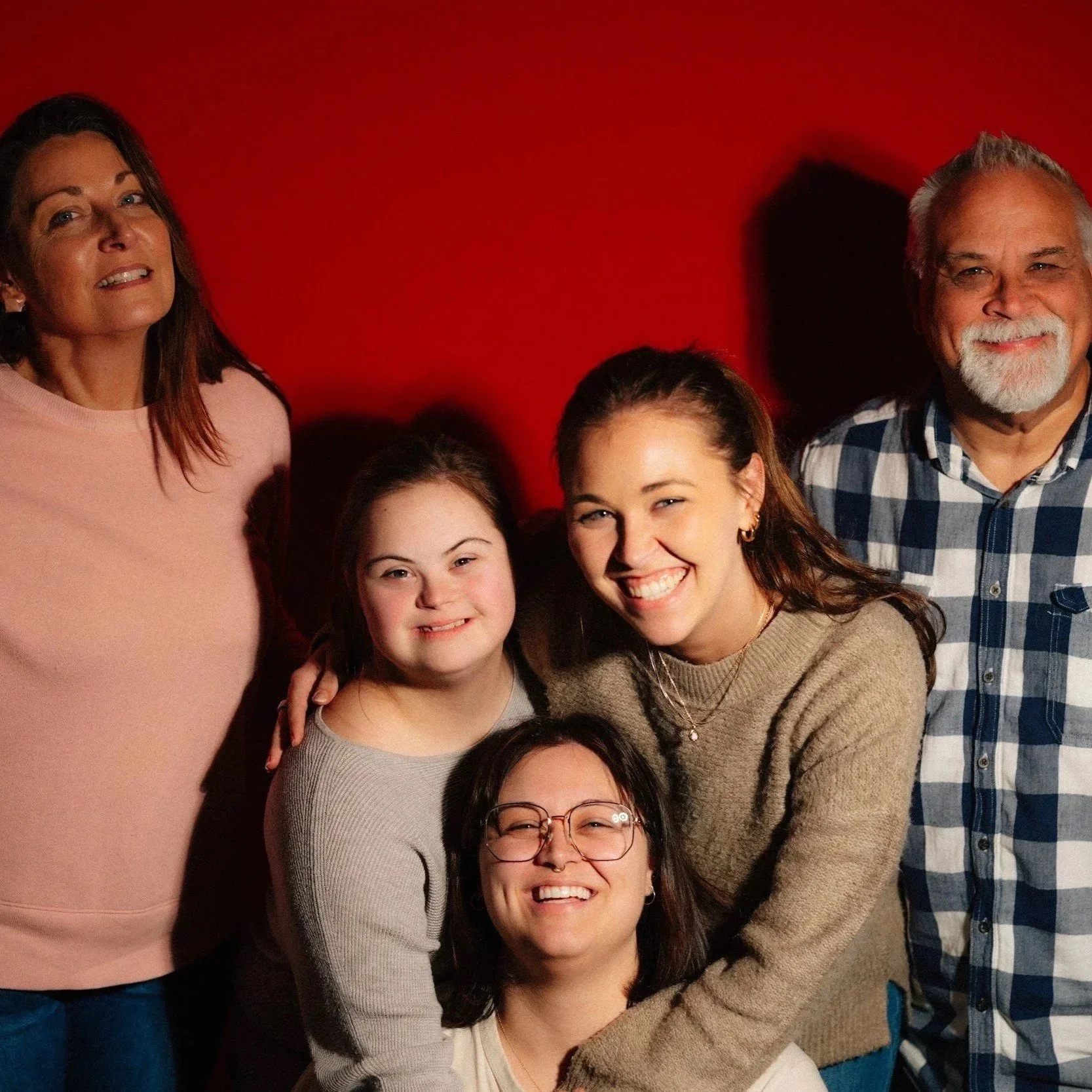 Group of six diverse people smiling and posing together against a red background, including women, men, and teenagers.