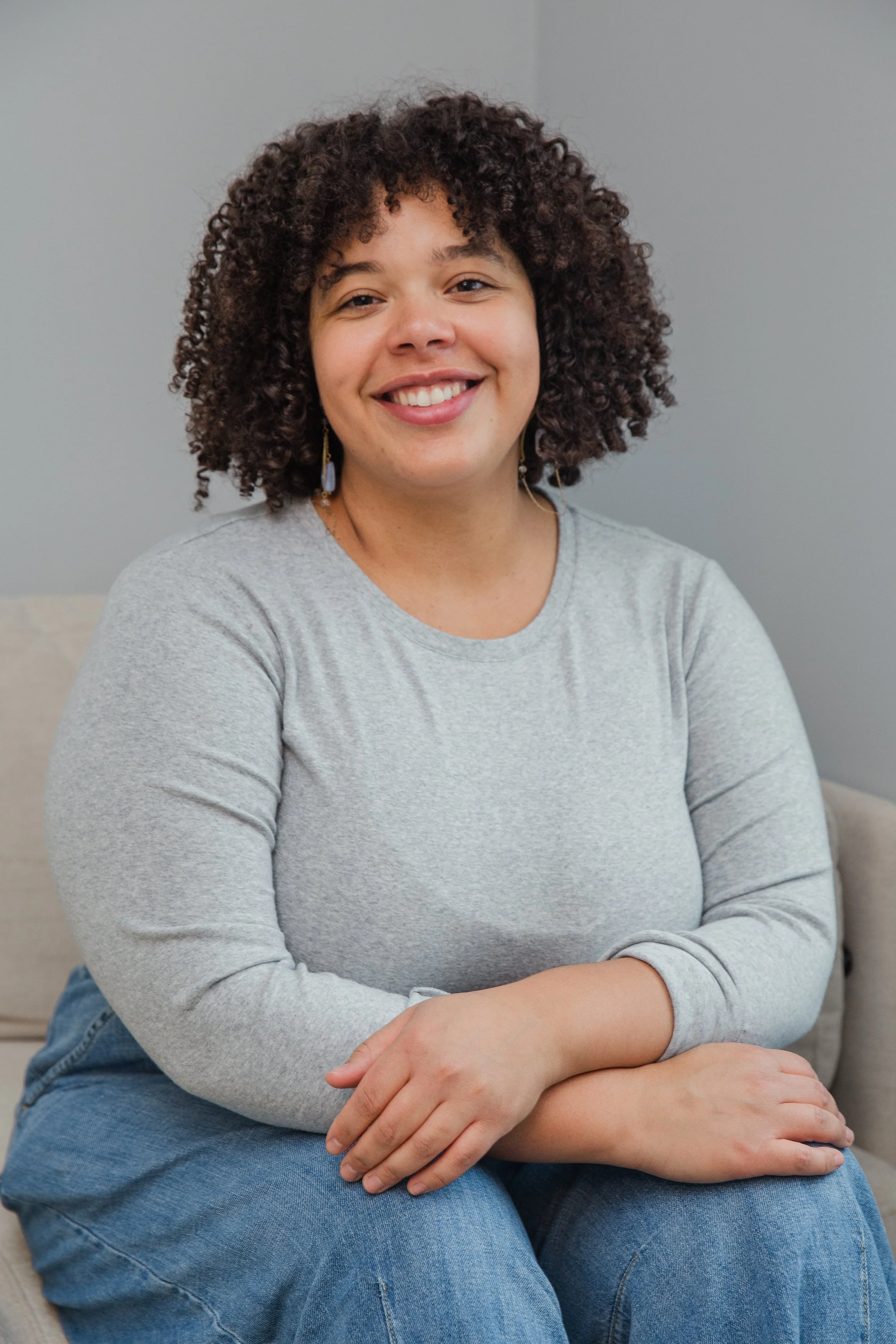 A woman with curly hair sitting on a couch, smiling, wearing a light gray long-sleeve shirt and blue jeans.