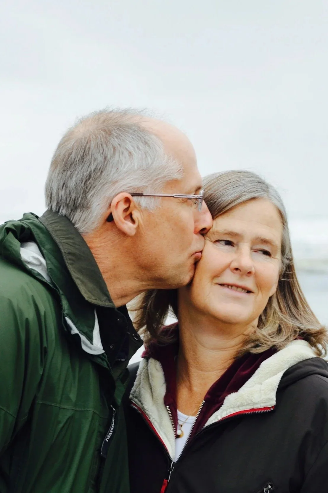 A man with gray hair and glasses is kissing a woman on the cheek. The woman has gray hair and is smiling slightly while looking off to the side. They are outdoors with a cloudy sky and water in the background.