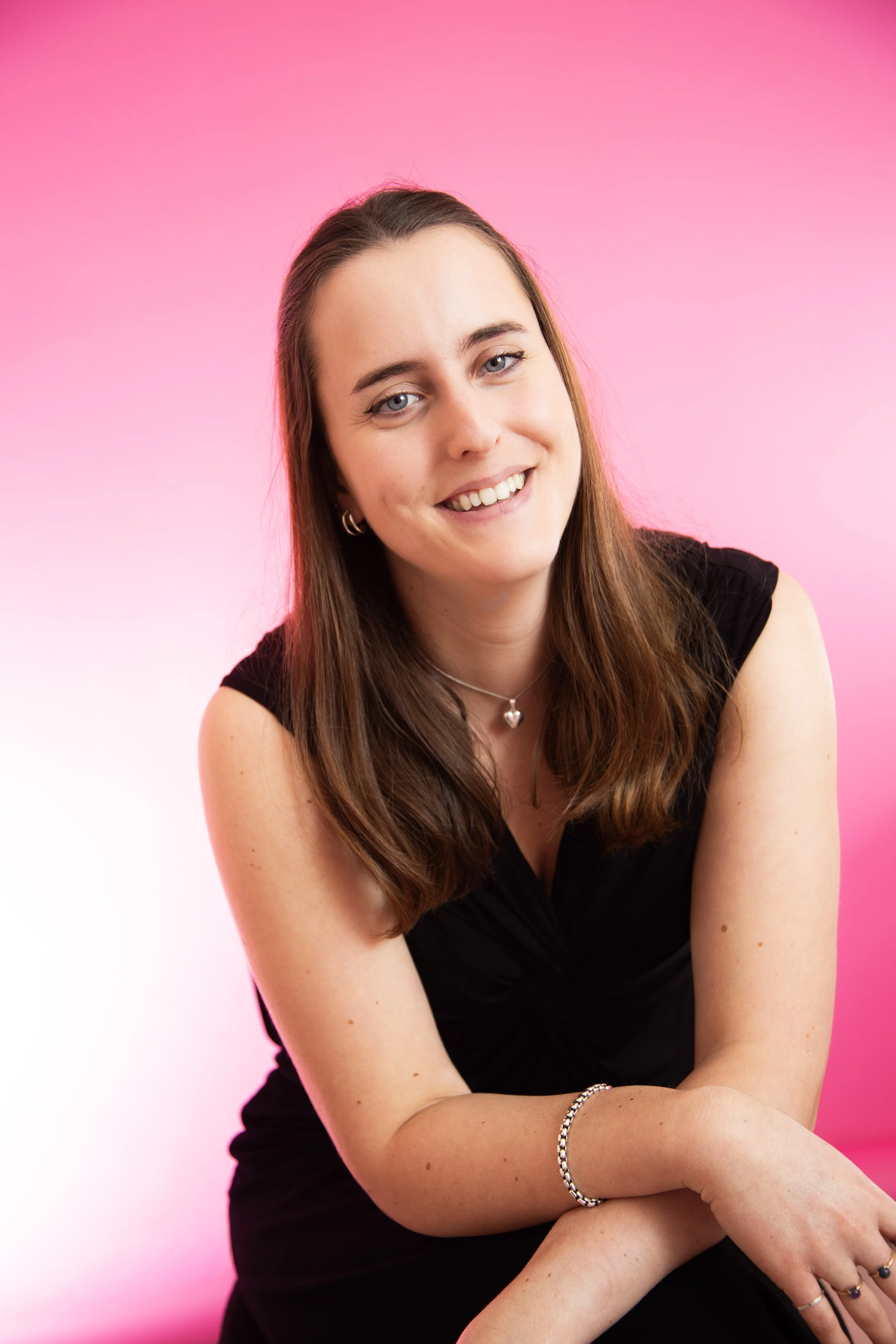 A young woman with long brown hair and blue eyes smiling while sitting against a pink background, wearing a black top, jewelry, and a silver bracelet.