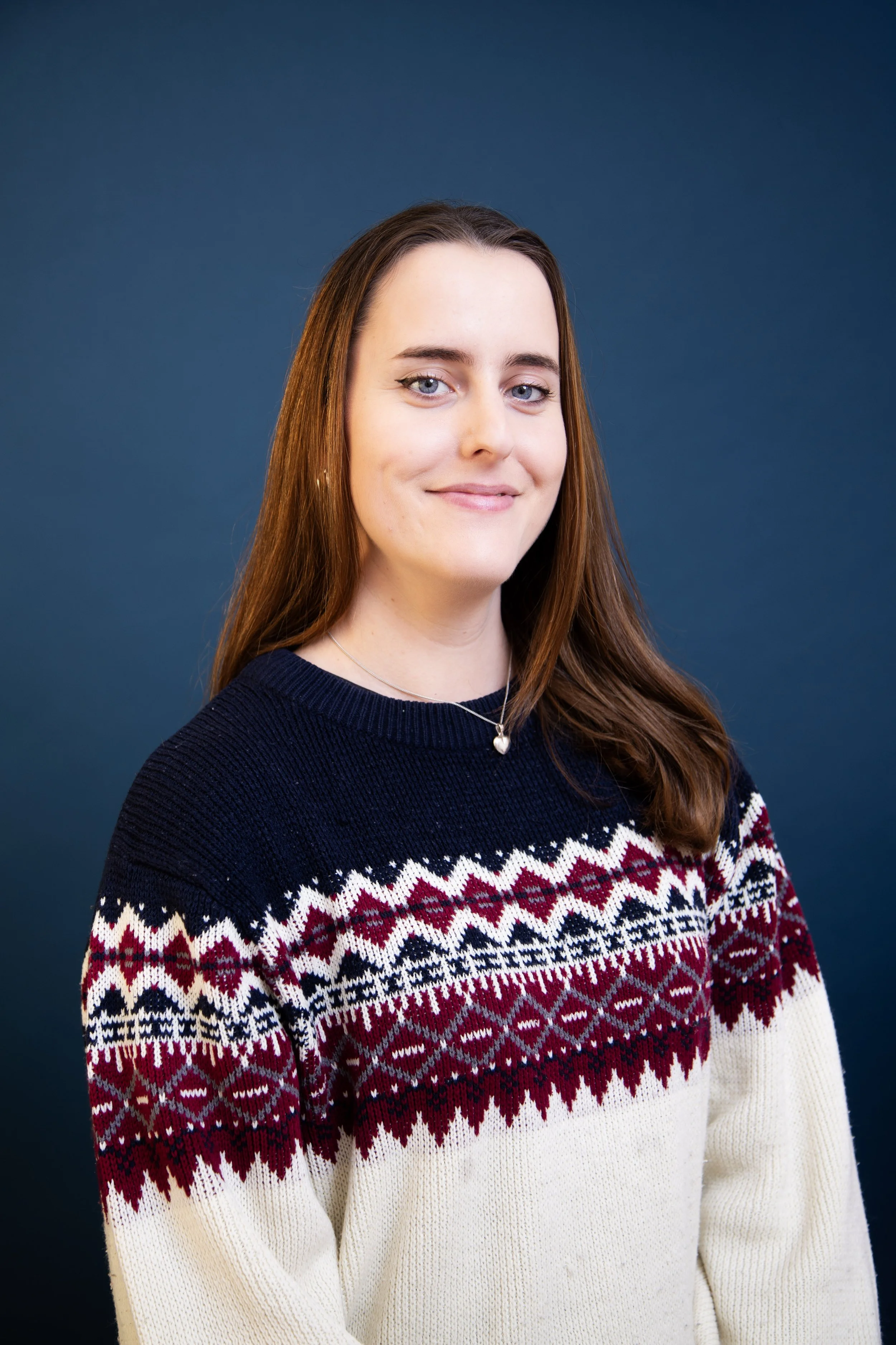 Young woman with long brown hair and blue eyes, wearing a navy blue sweater with a red, white, and navy patterned design, and a silver heart pendant necklace, standing against a dark teal background.